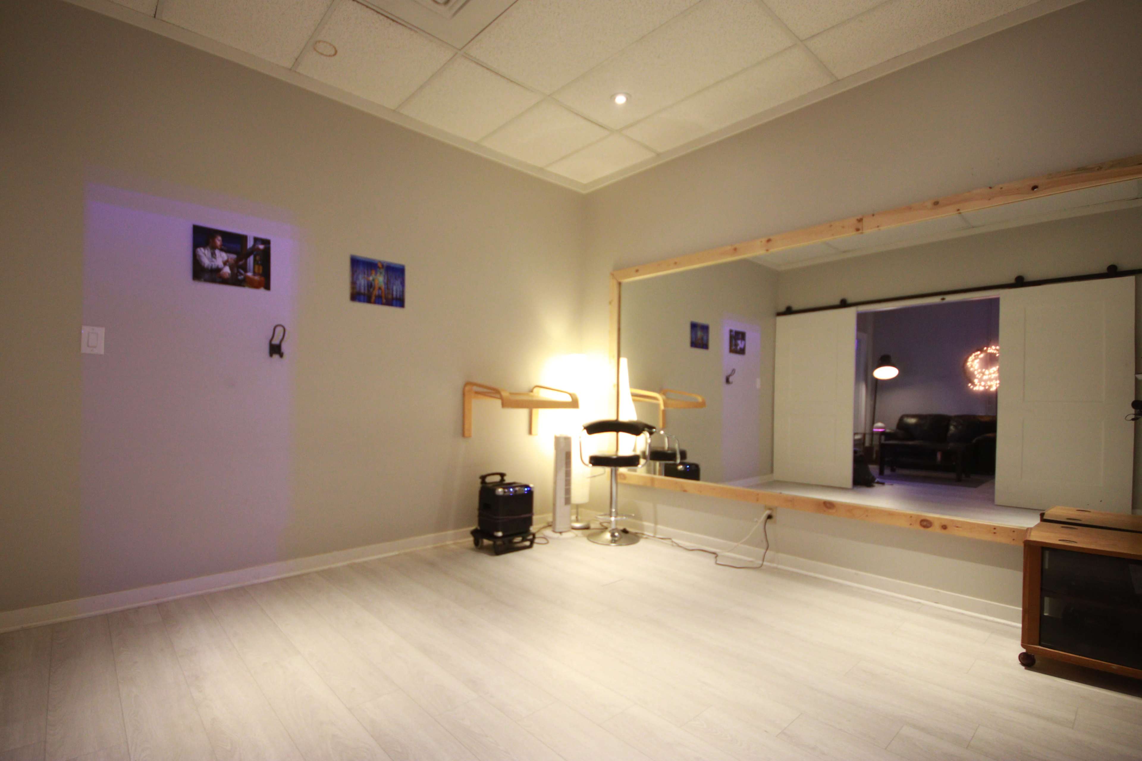 The image shows a minimalistic room with a large mirror, a chair, and faint lighting, featuring light-colored walls and wooden accents.