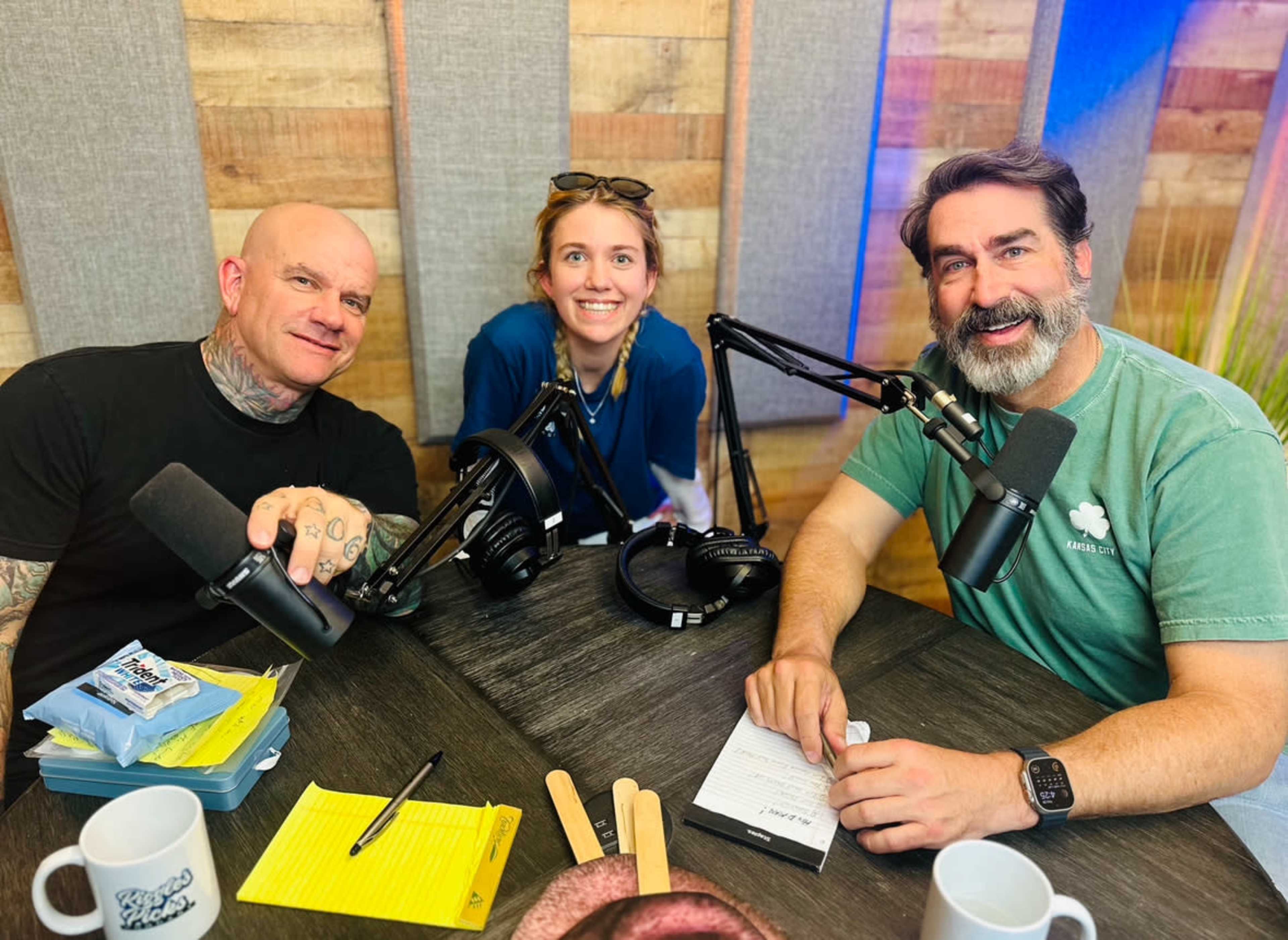 Three individuals sit at a table with microphones and recording equipment, surrounded by wooden paneling.