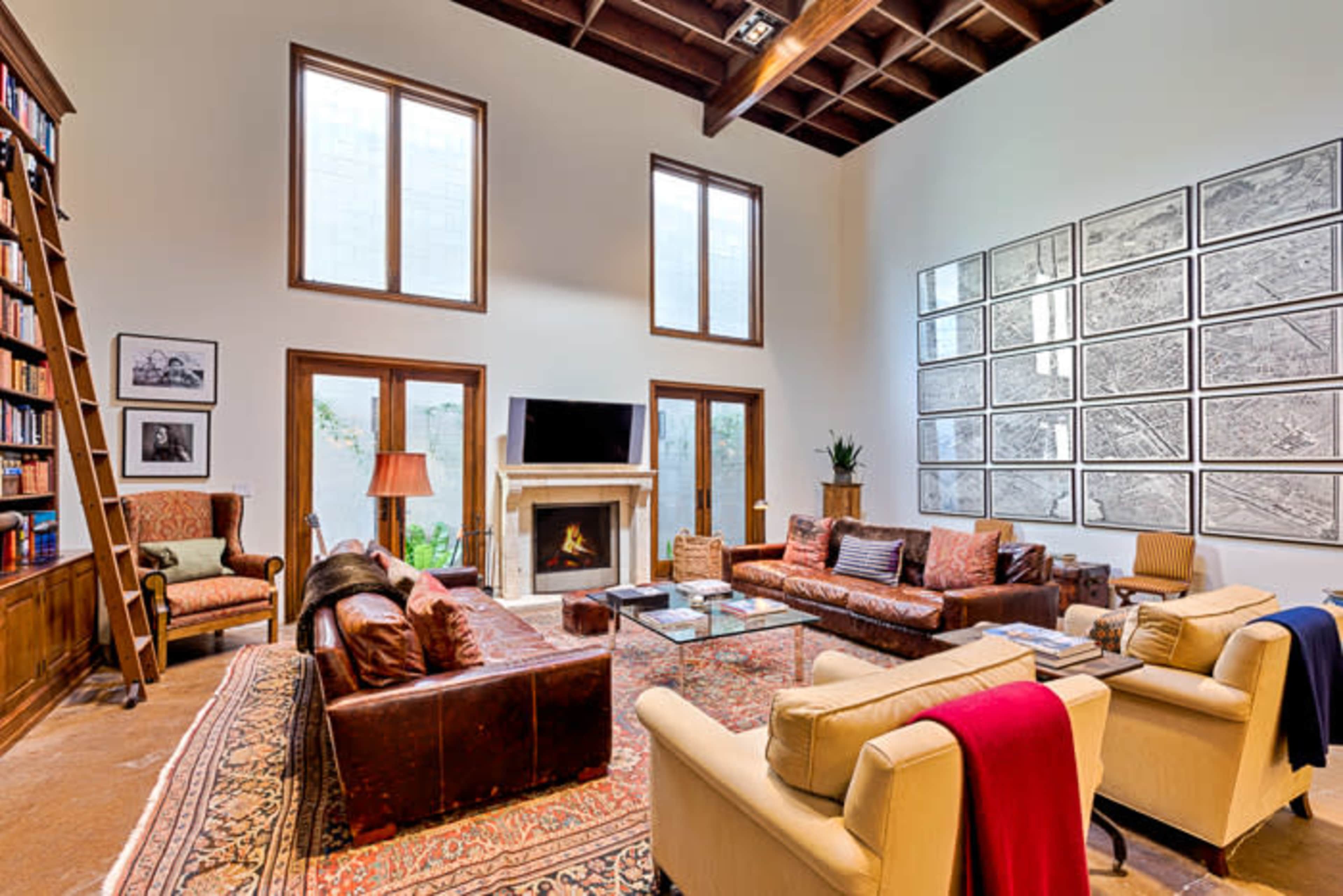 The image shows a spacious living room with high ceilings, a fireplace, a large bookshelf, and various seating arrangements, including leather couches and armchairs.
