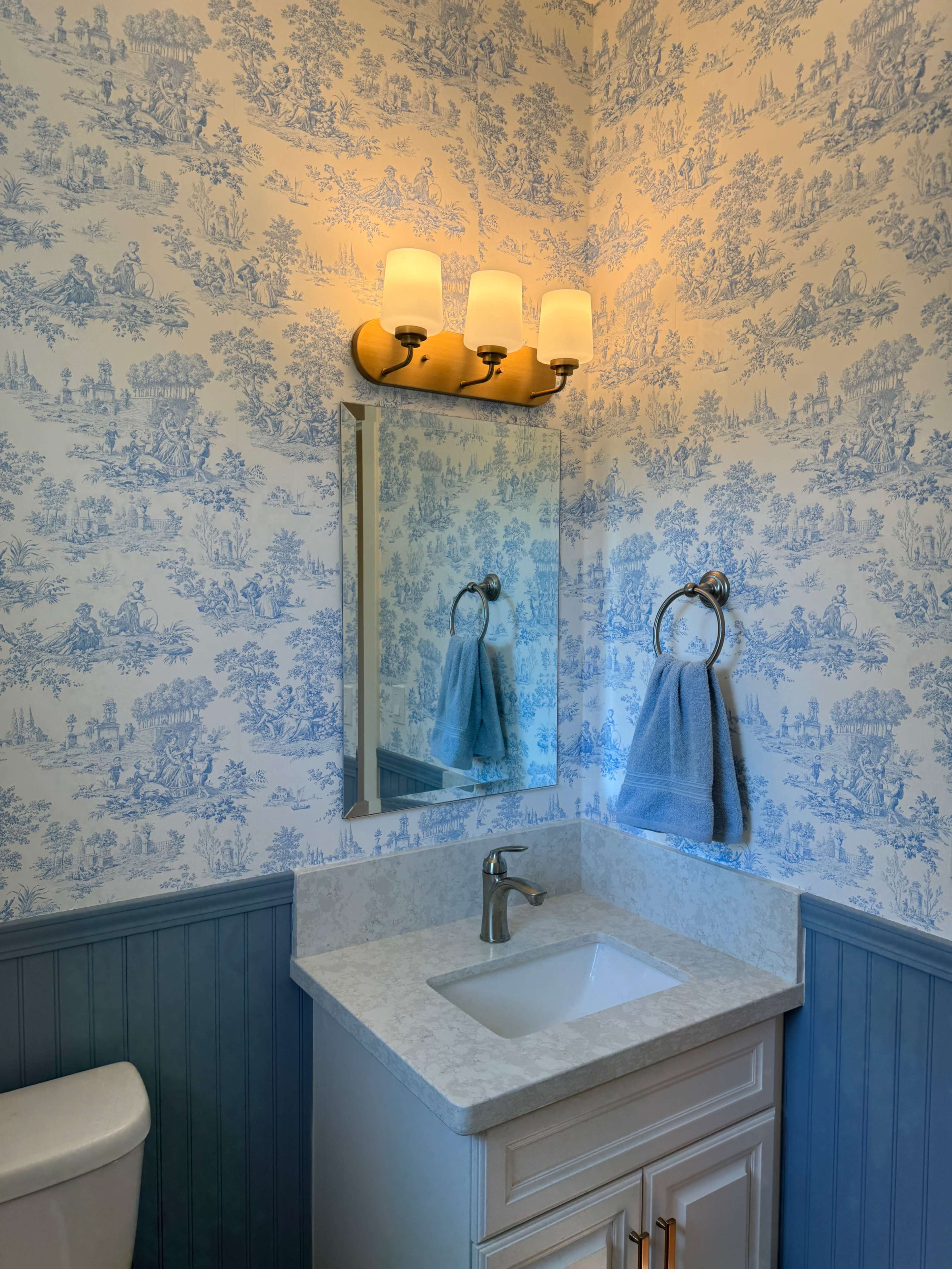 The image features a bathroom with blue toile wallpaper, a white marble countertop with a sink, a mirror, a light fixture above, and a towel hanging on a silver ring.