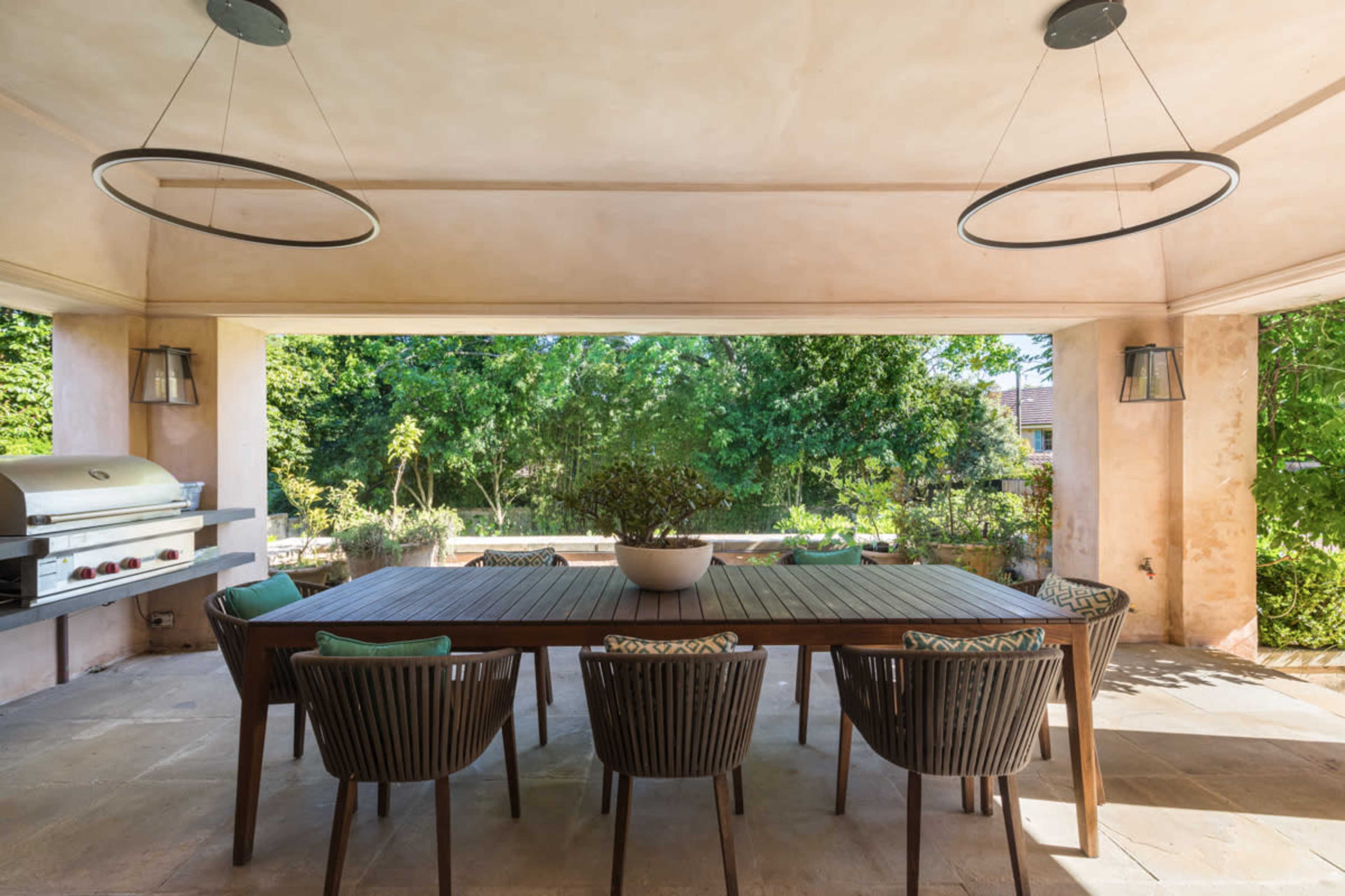 A modern outdoor dining area features a large wooden table surrounded by chairs, with a stainless steel grill visible in the background and lush greenery outside.