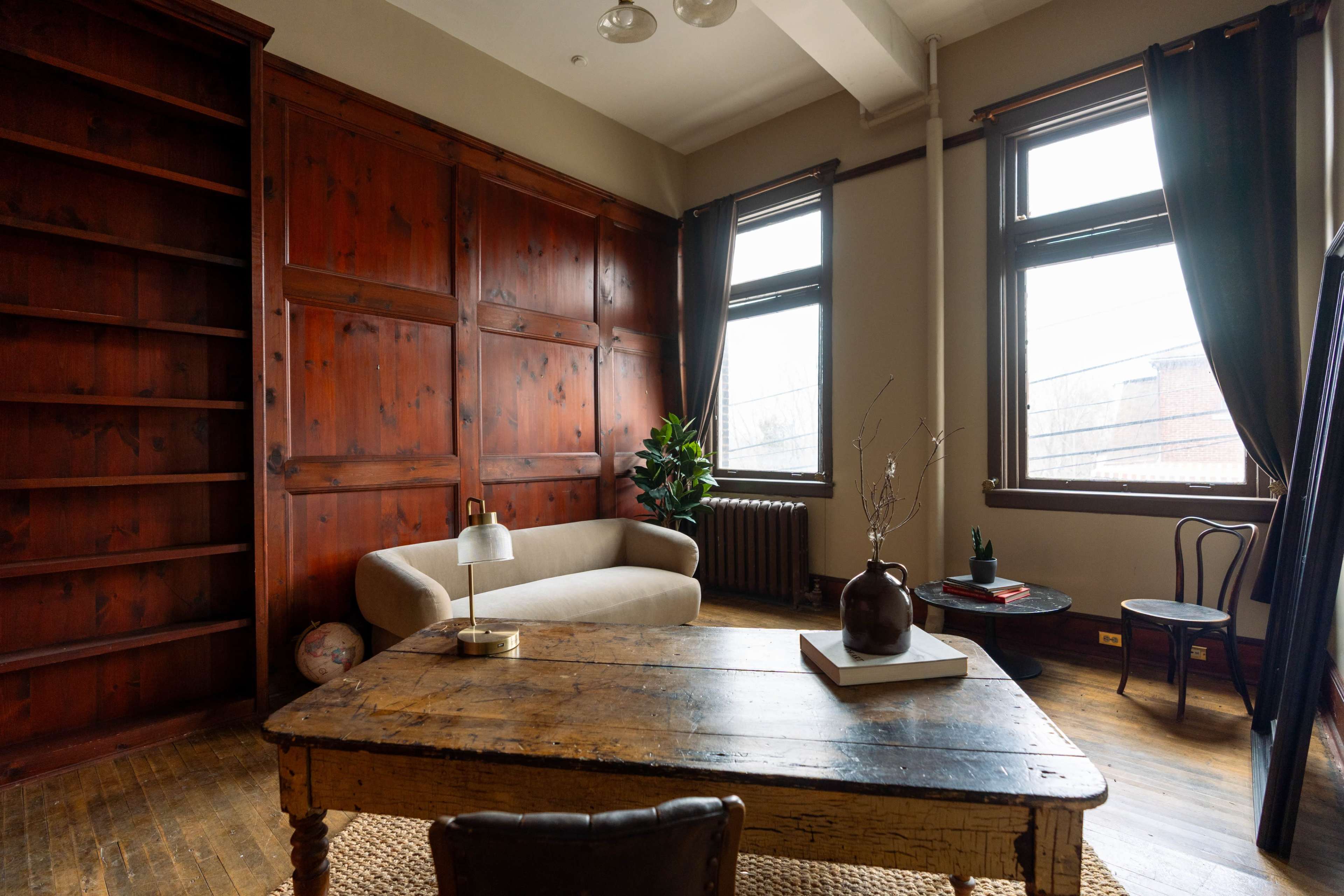 The image depicts a cozy interior of a room featuring wooden walls, a simple sofa, a wooden table, and a few decorative elements along with large windows allowing natural light.