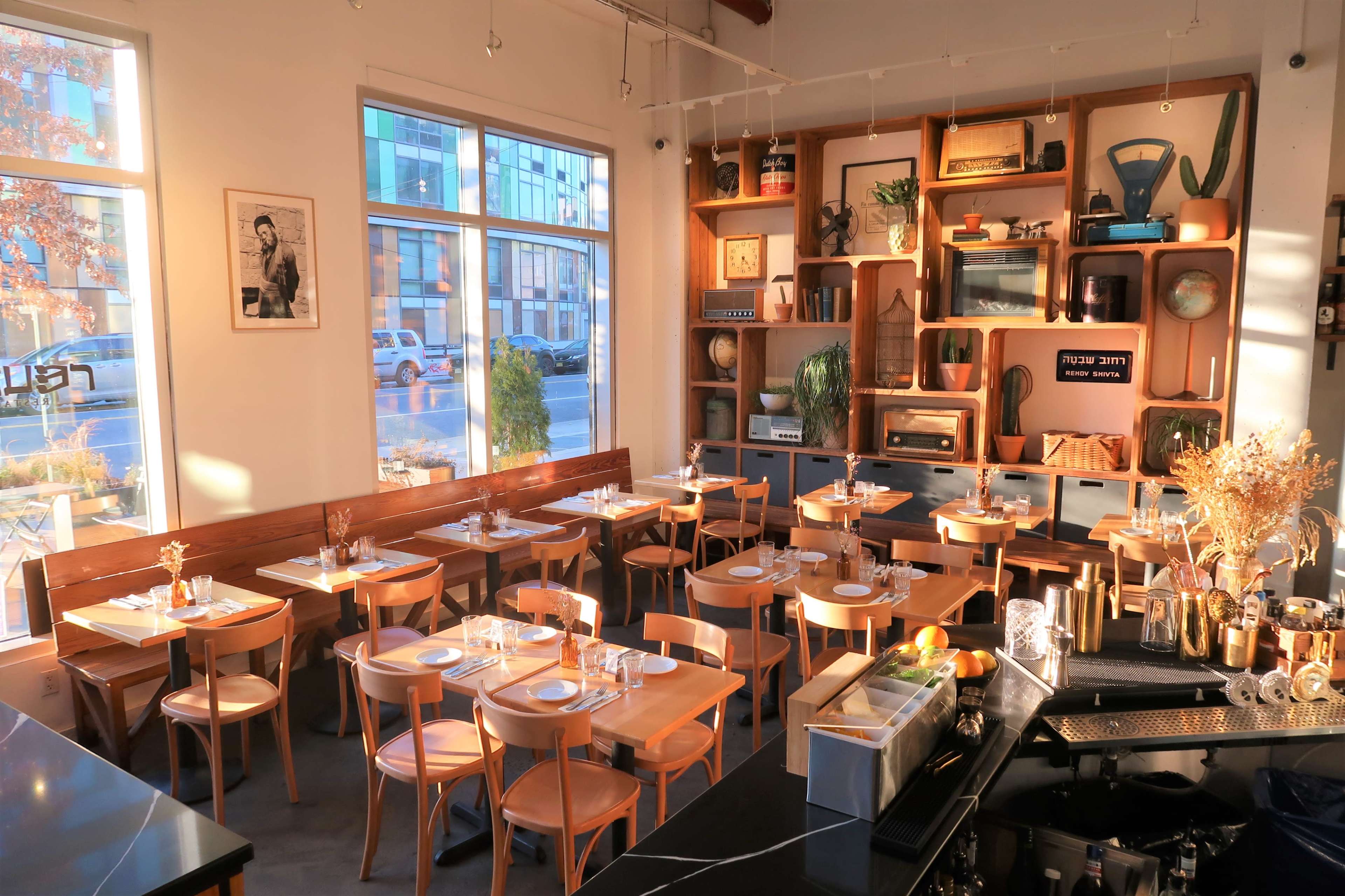 A modern restaurant interior with wooden tables, chairs, and a decorative shelving unit, illuminated by natural light through large windows.
