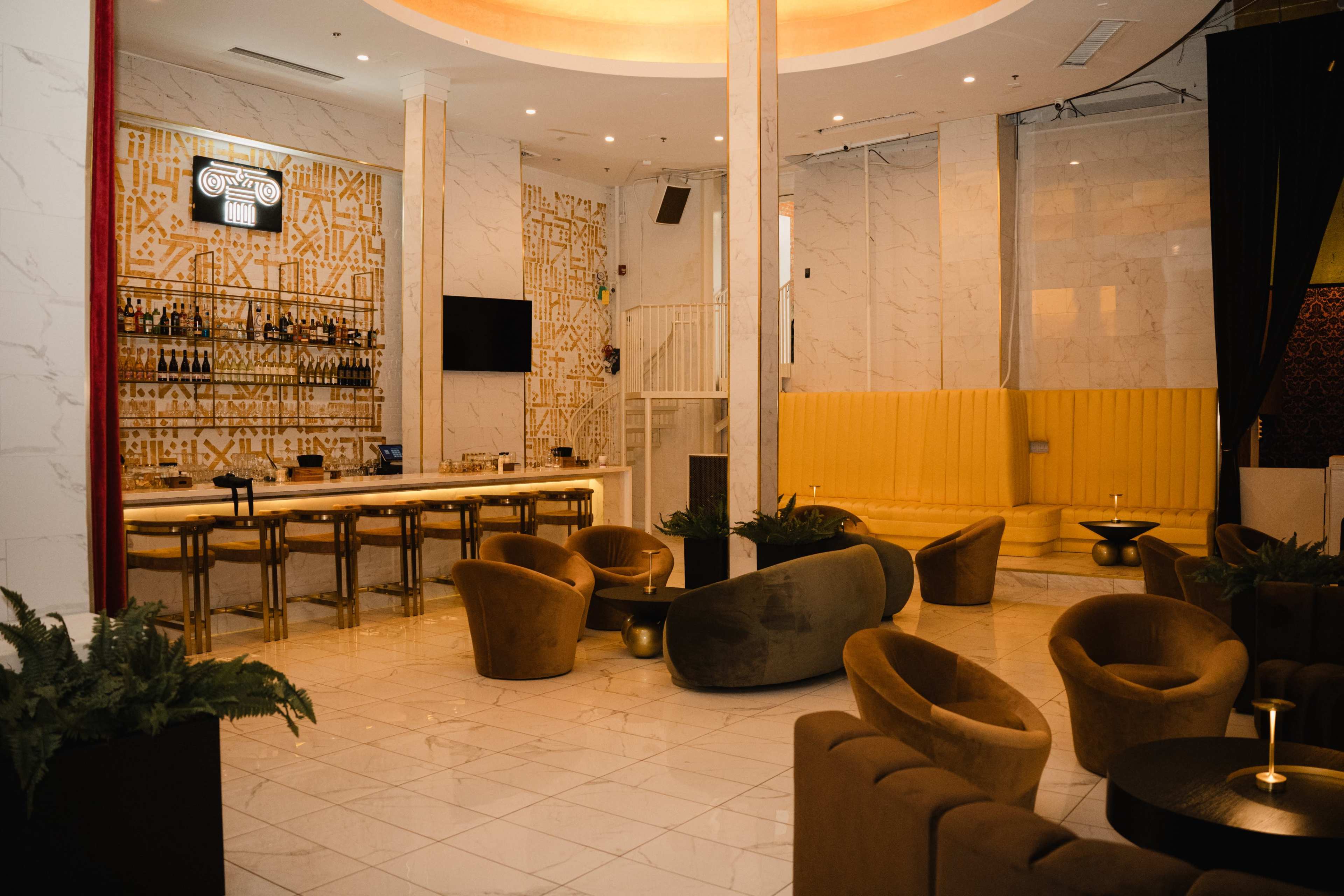 The image shows a modern lounge area featuring a bar with high stools, a large yellow booth, and stylish seating arrangements.