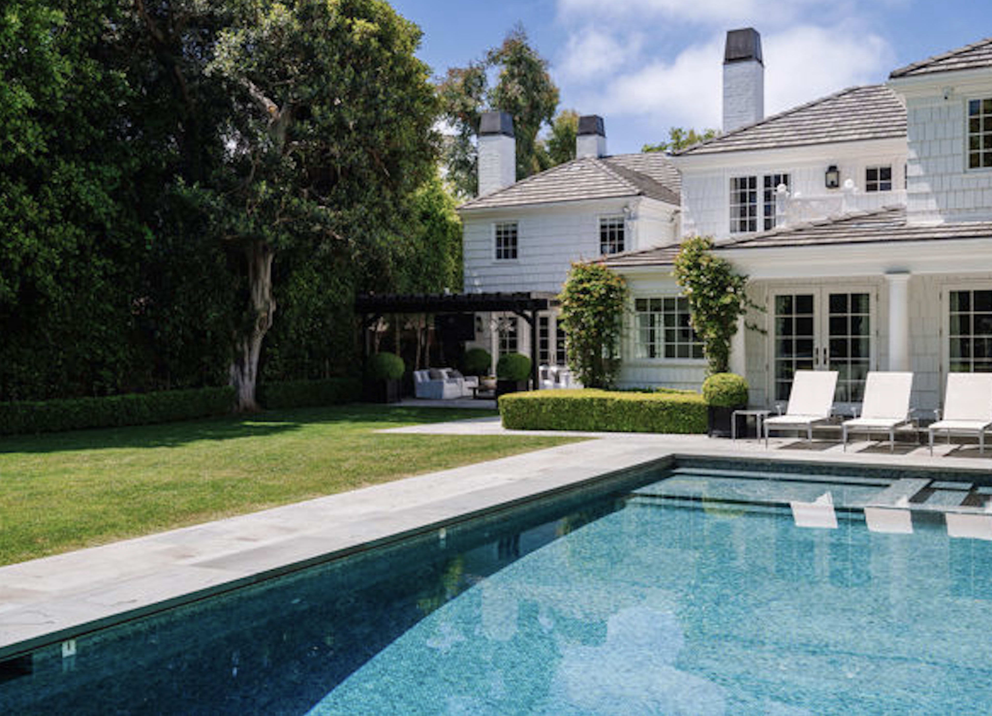 The image shows a modern white house with a well-maintained lawn and a swimming pool surrounded by lounge chairs.