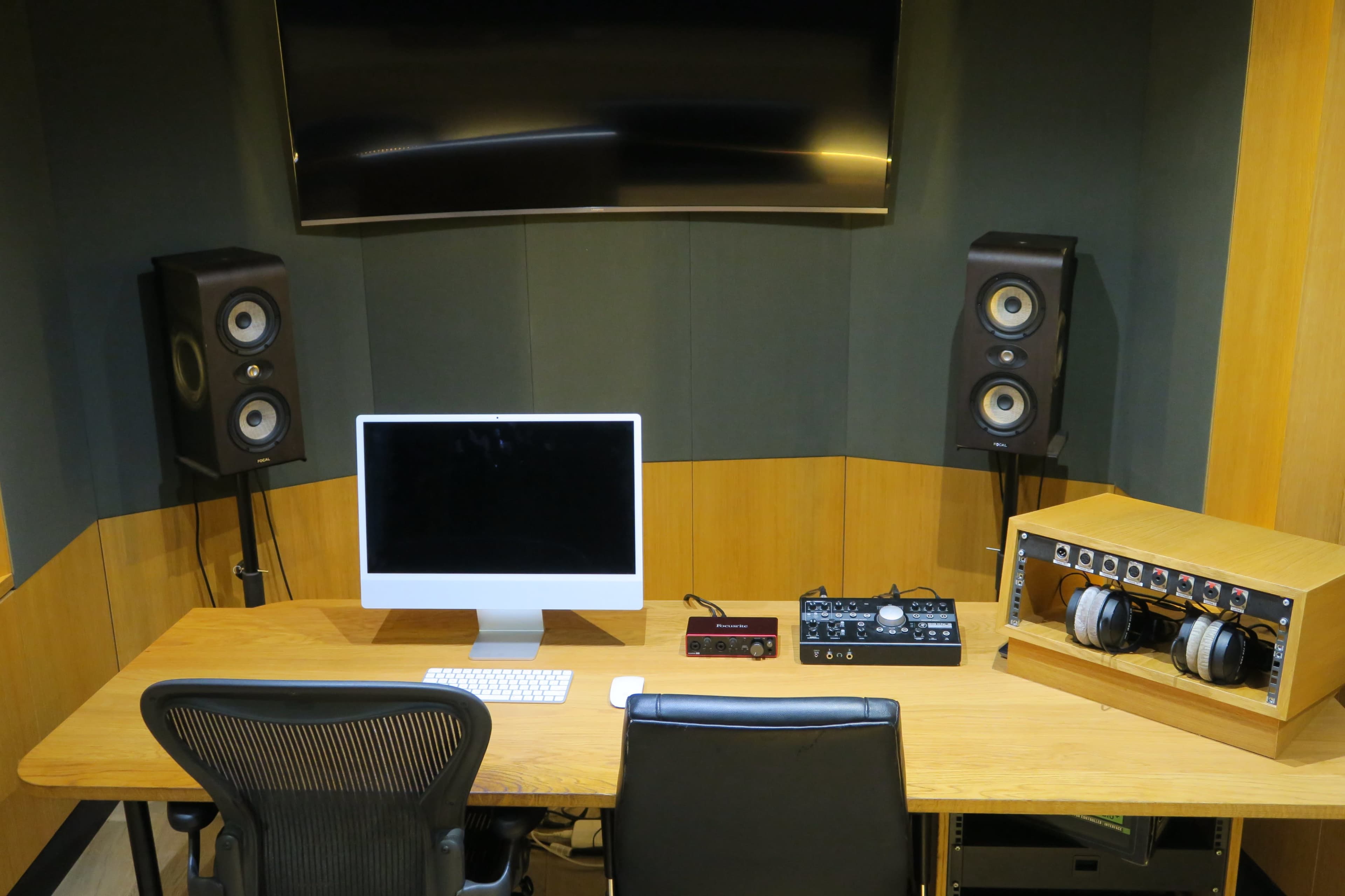 The image shows a compact audio recording studio featuring a computer with a large monitor, studio speakers on either side, a digital audio interface, and a headphone station on a wooden desk.