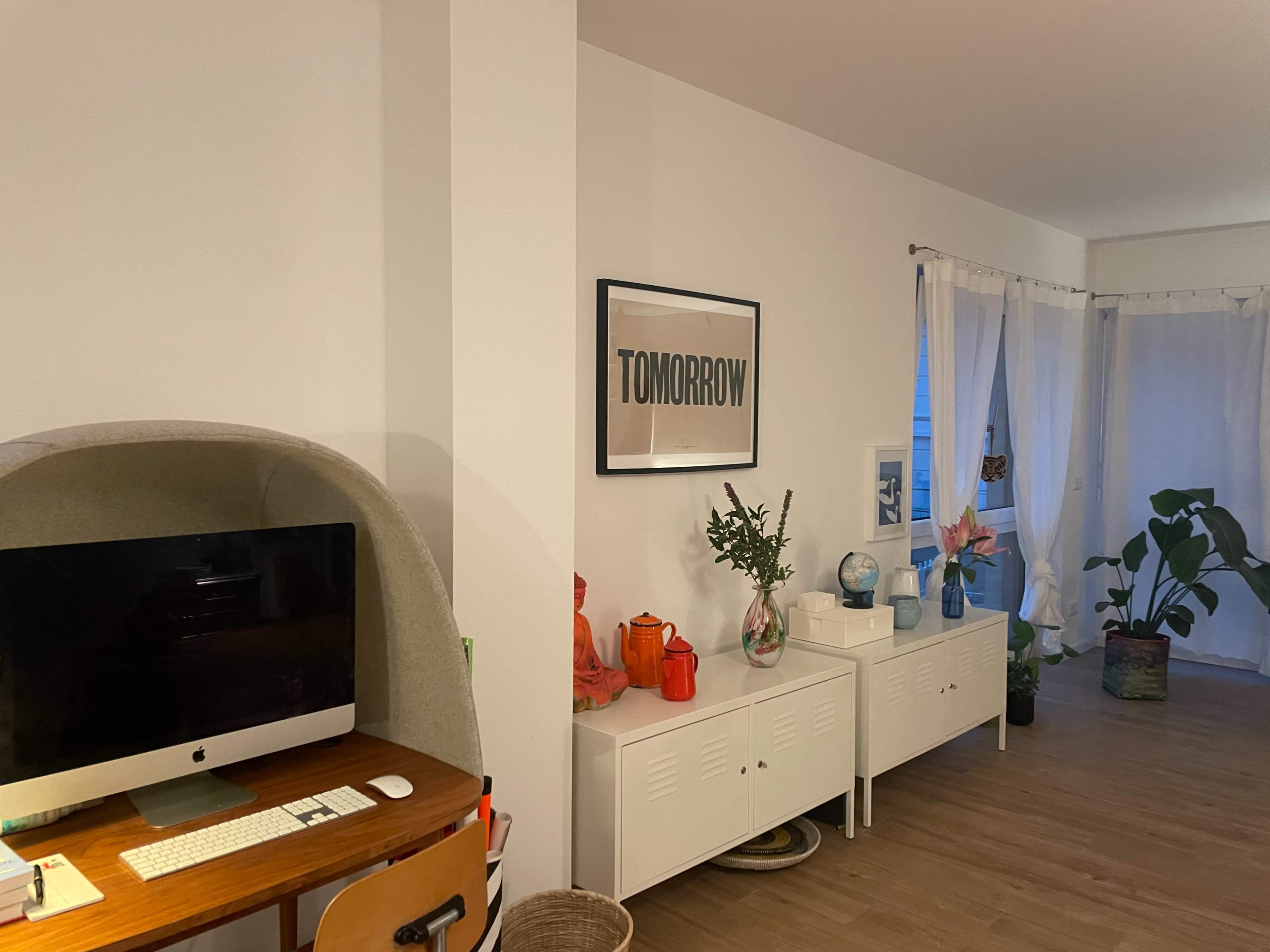 A modern living space features a desk with a computer, a white cabinet displaying decorative items, and a framed poster that reads "TOMORROW" on the wall.