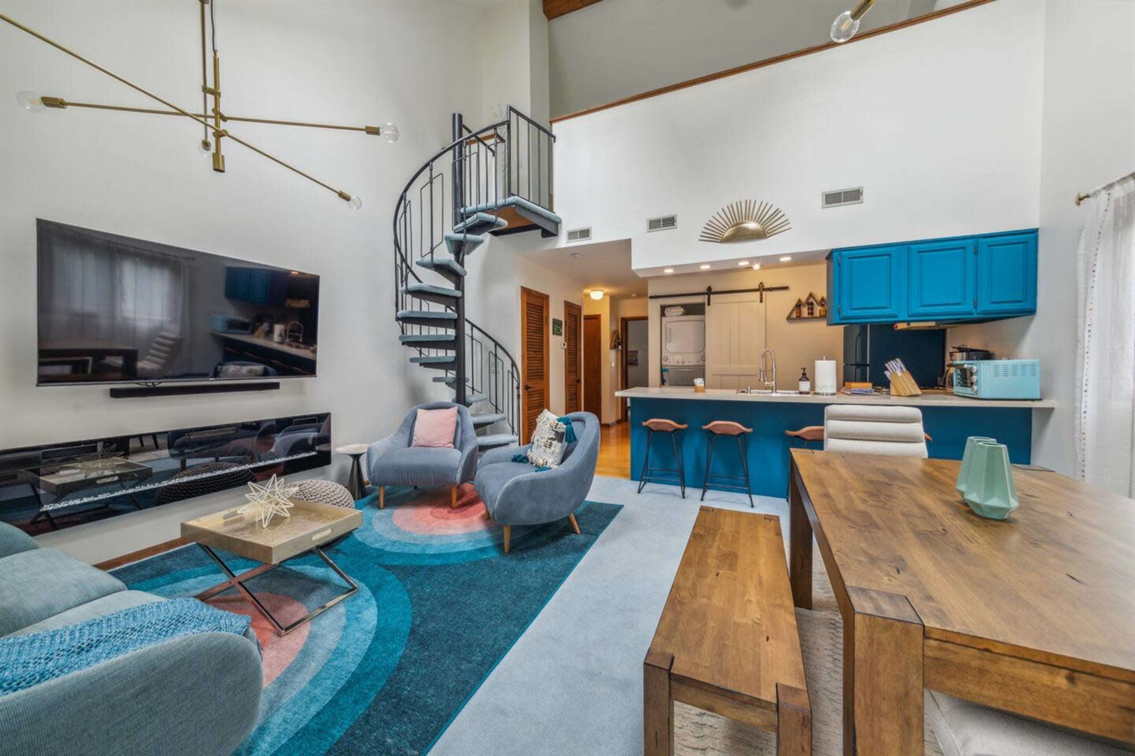 A modern living room with a spiral staircase leading to an upper level, featuring a large television, a blue kitchen area, and contemporary furniture arranged around a colorful area rug.