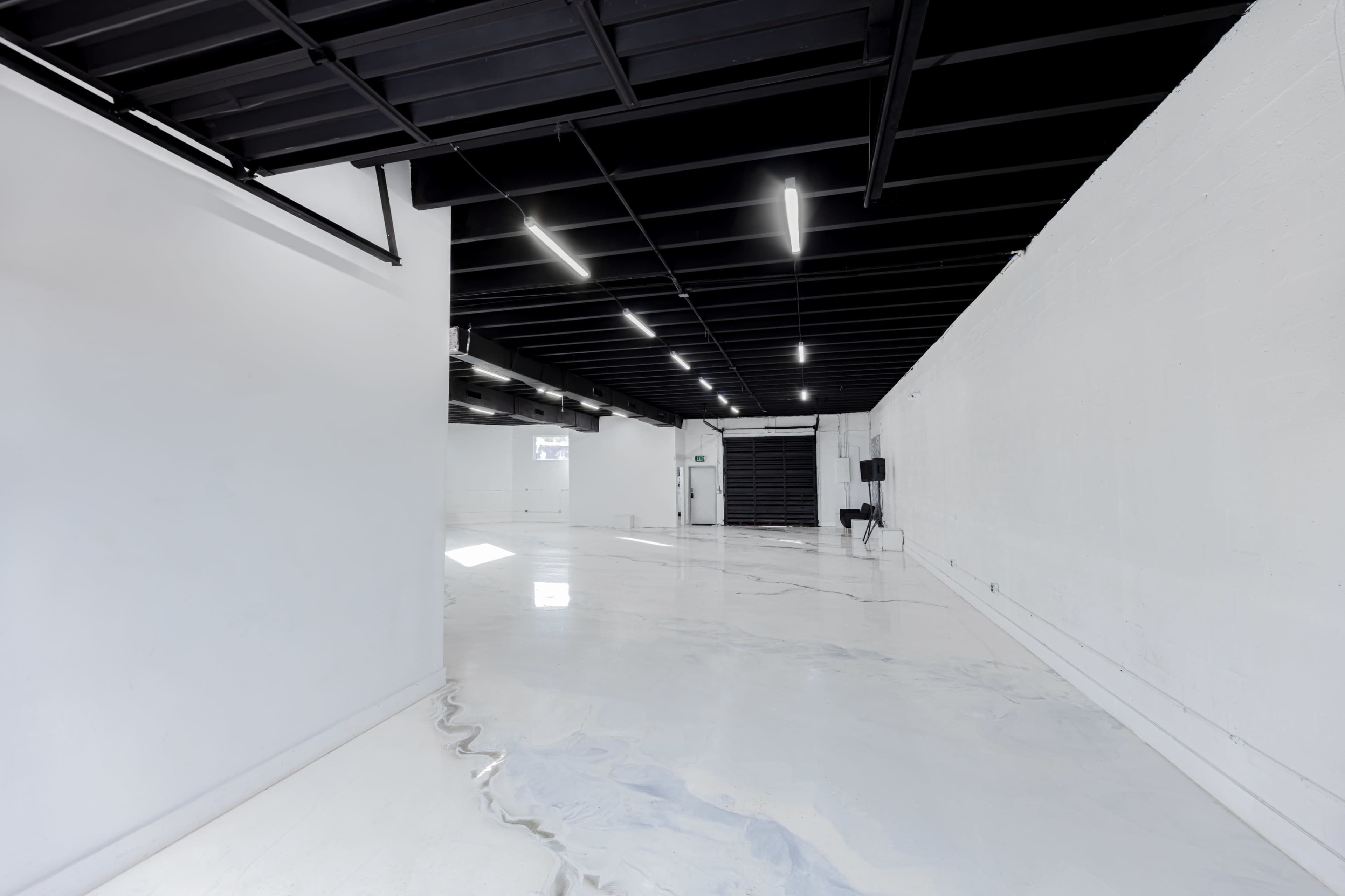 The image shows a spacious, empty room with white walls and a glossy floor, illuminated by fluorescent lighting.