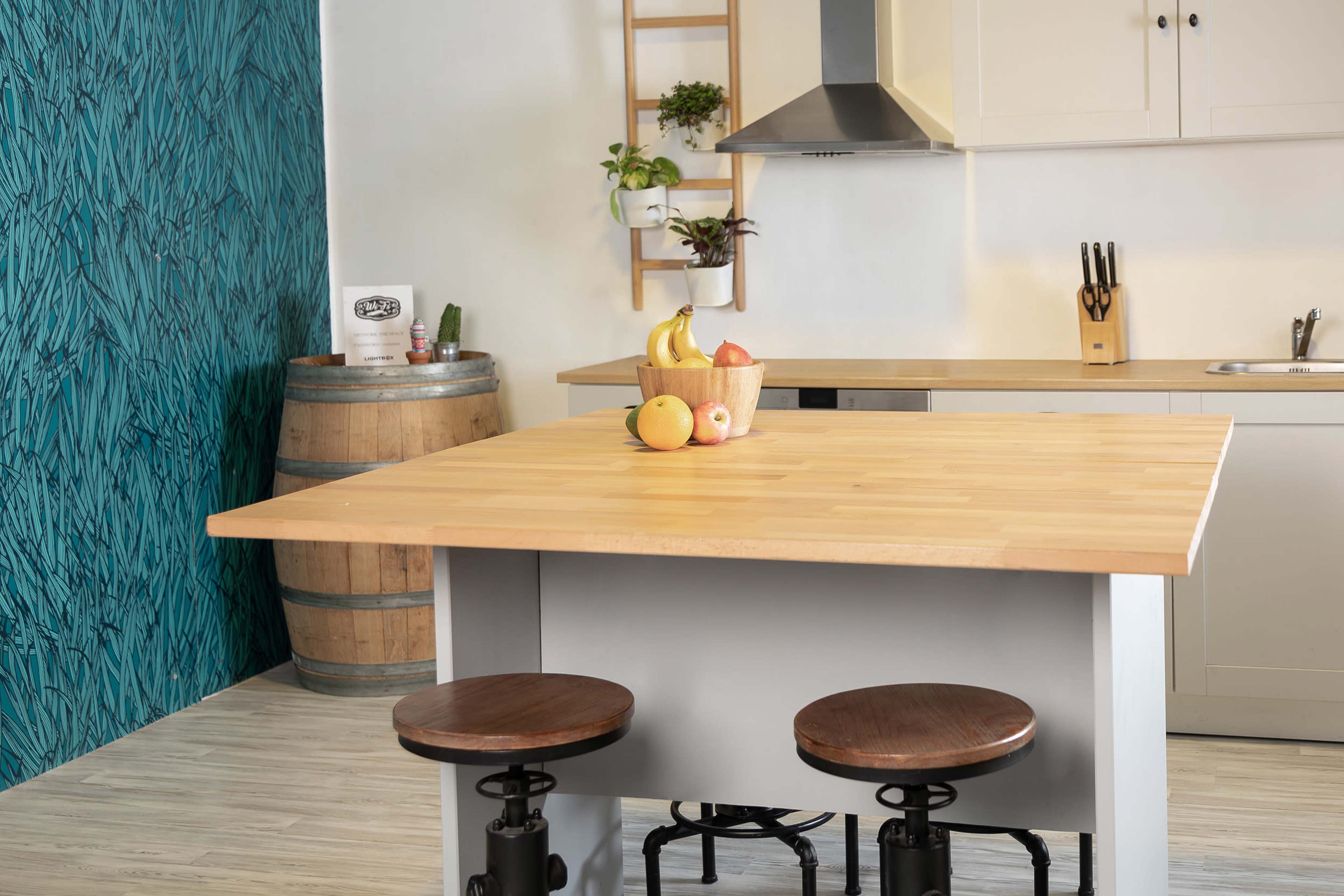 A modern kitchen features a wooden island with two stools, a fruit bowl, and a decorative wall with green patterns.