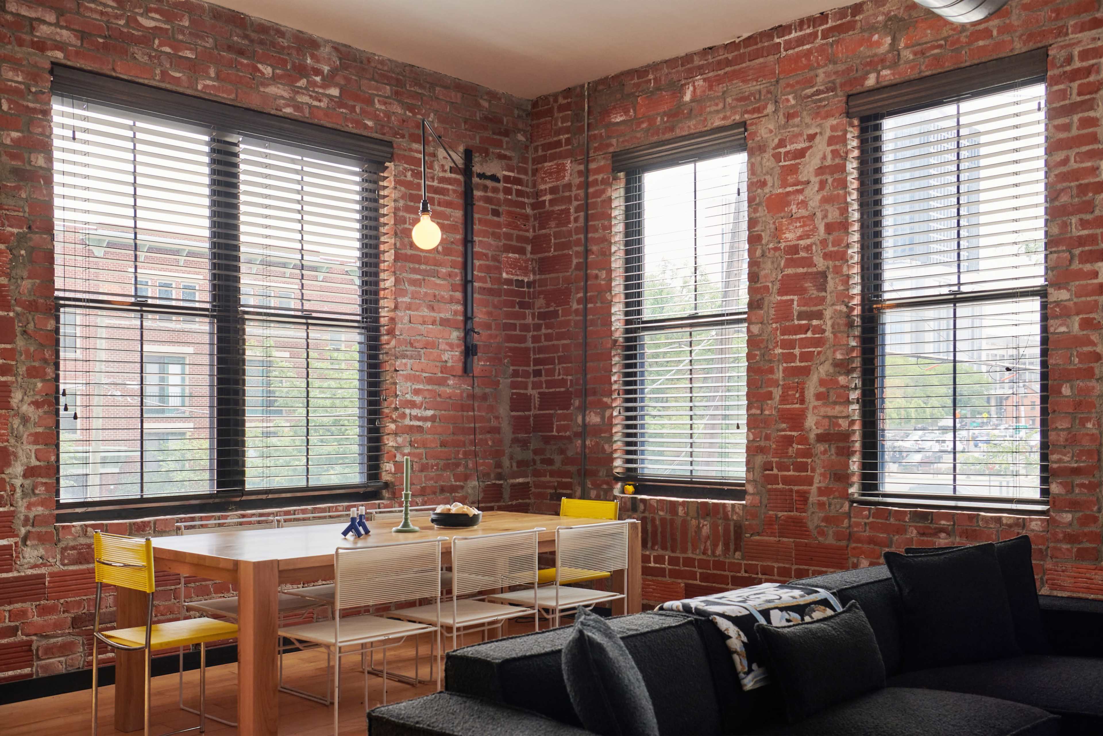 A dining table with yellow and clear chairs is positioned near large windows in a room with exposed brick walls.