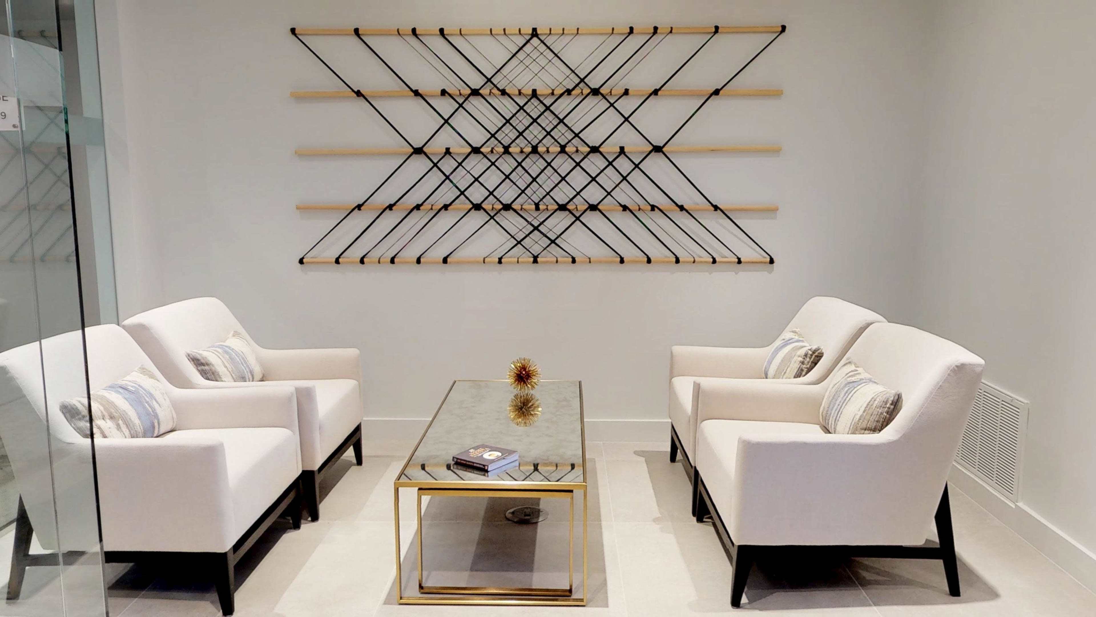 The image shows a modern seating area with two beige chairs facing a glass coffee table, set against a minimalist wall adorned with a geometric art installation.