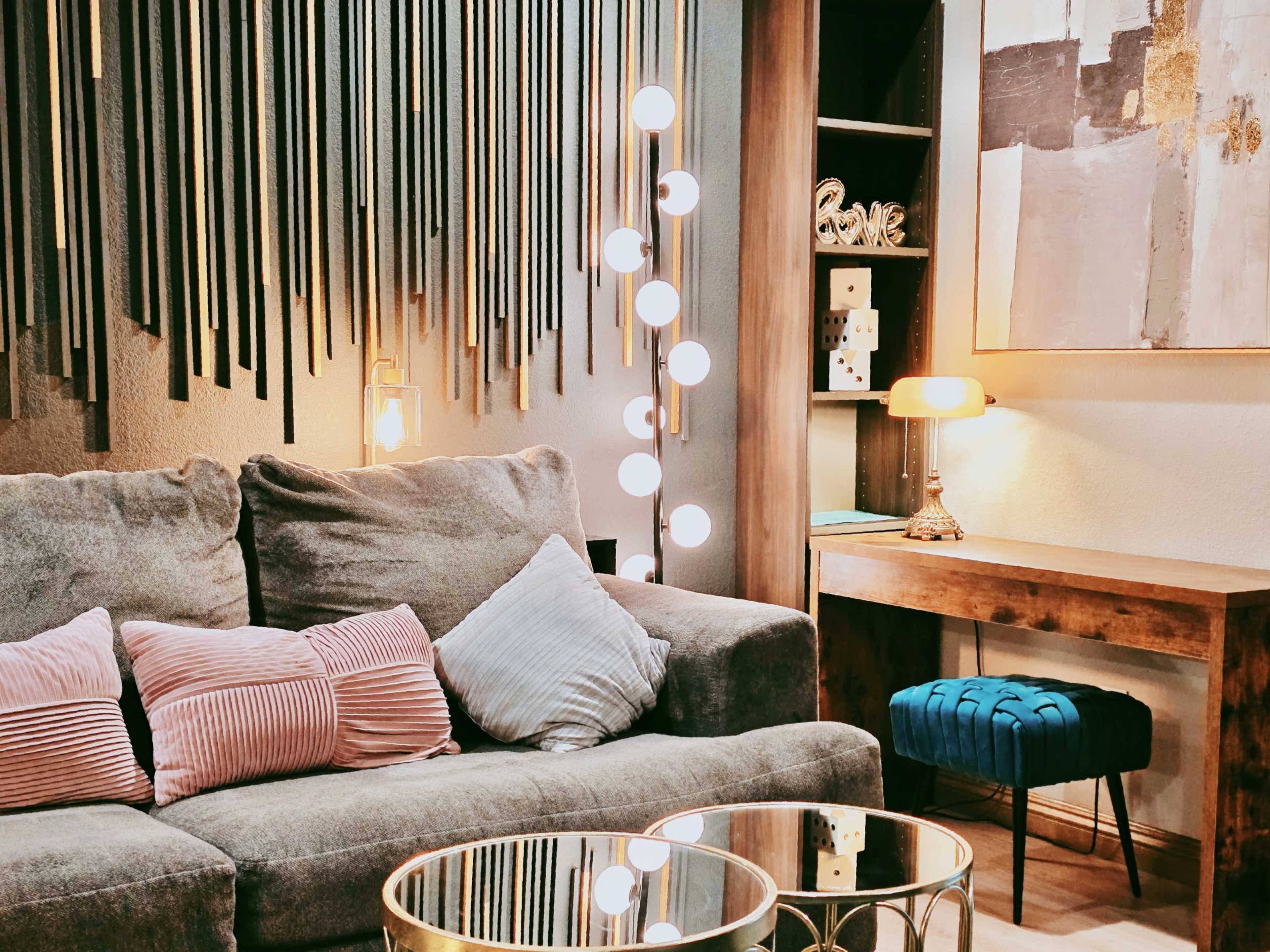A gray sofa with several decorative pillows sits in a stylish living room featuring a lamp and a wooden console table against a backdrop of vertical wooden slats.