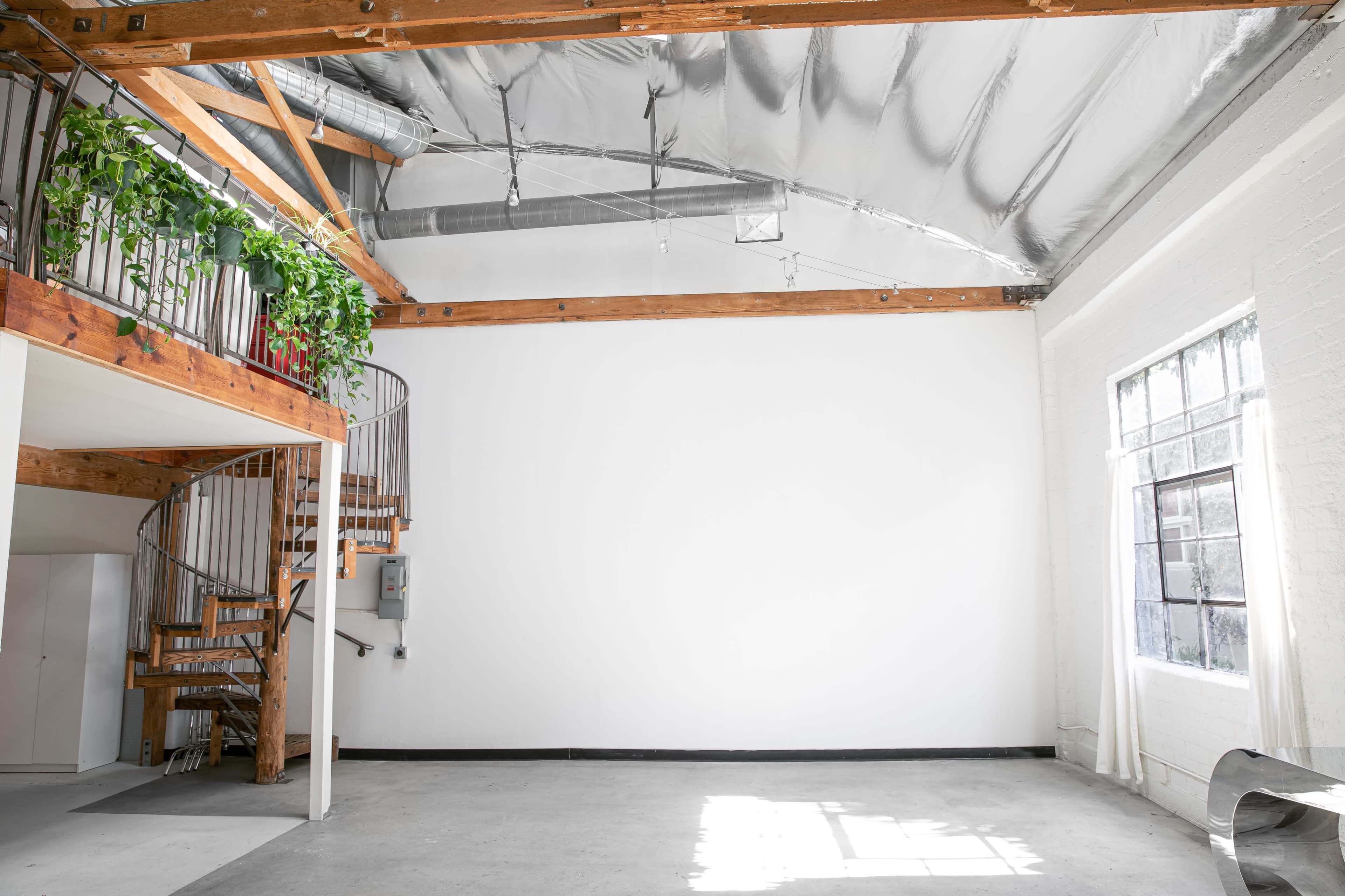 An industrial-style room with a spiral staircase, wooden beams, and a large window allowing natural light to enter.