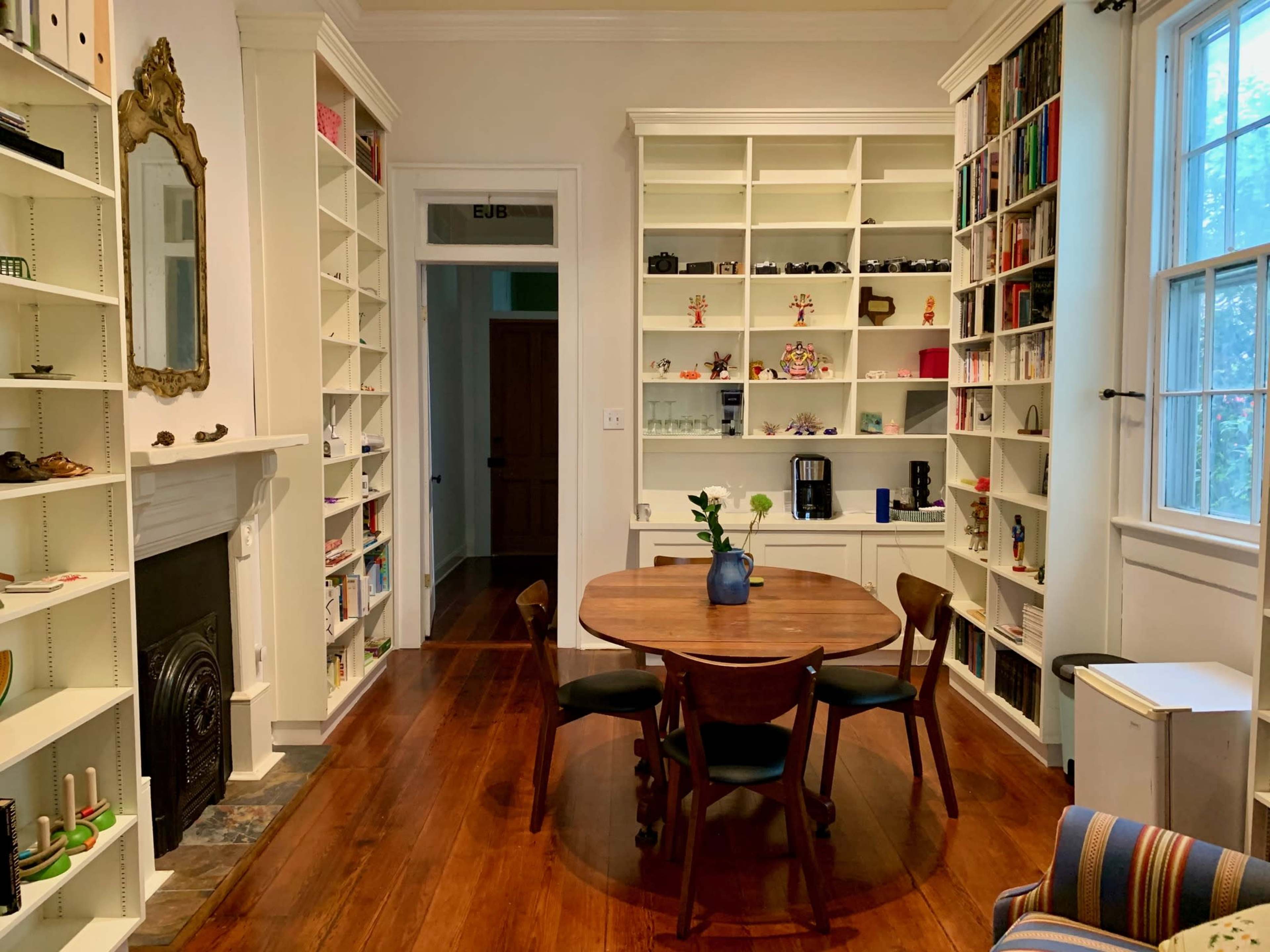 The image shows a cozy room with a round wooden dining table surrounded by three black chairs, flanked by white bookshelves filled with various items and an open doorway leading to another room.