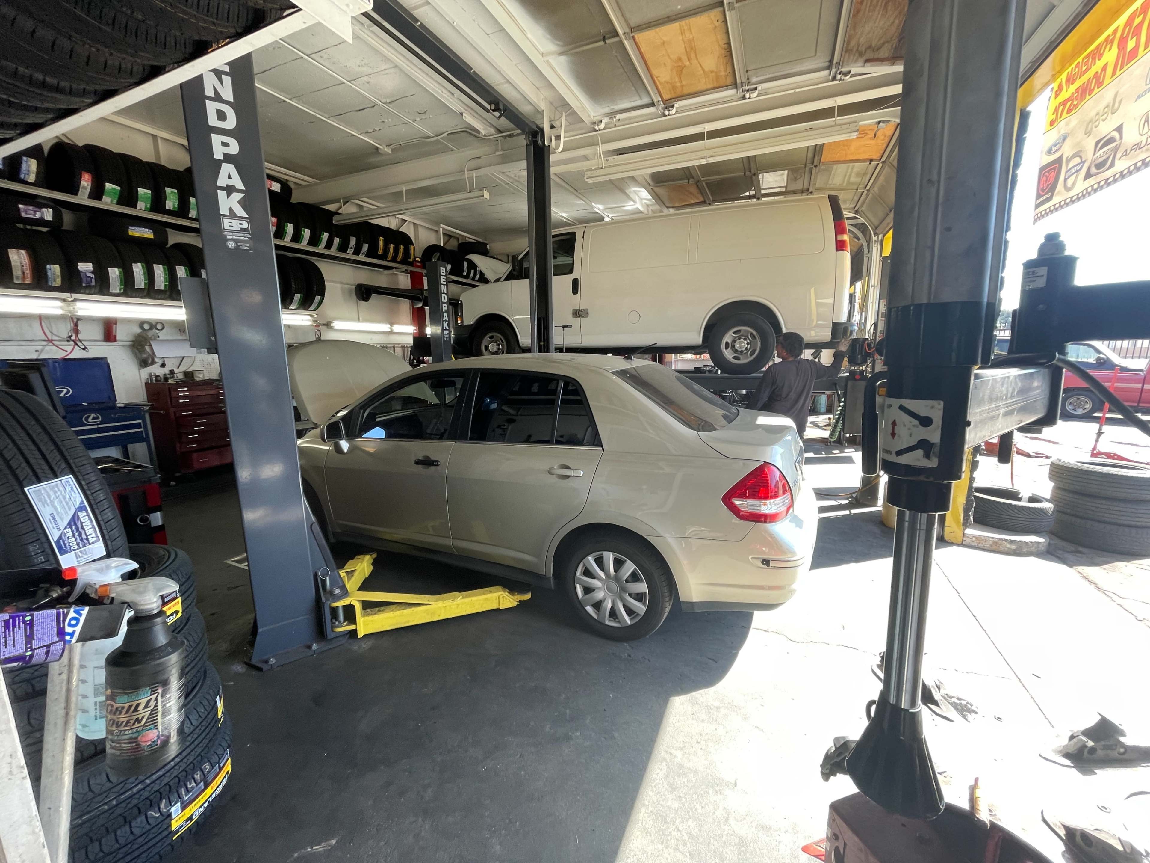 A light-colored sedan is being serviced on a lift inside a tire shop, while a van is elevated on another lift nearby.