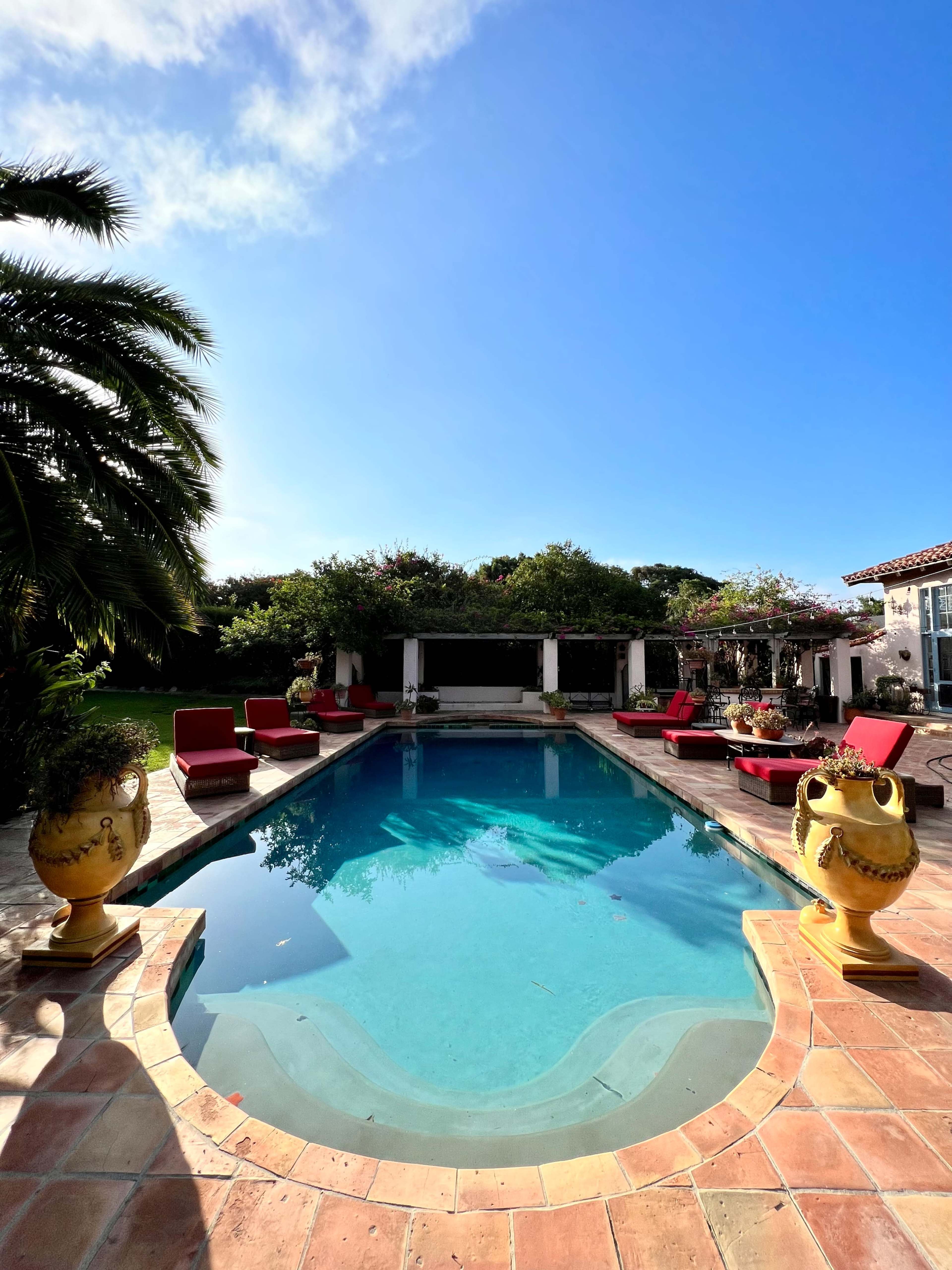 A clear swimming pool surrounded by red lounge chairs and ornate yellow planters, with a lush garden in the background.