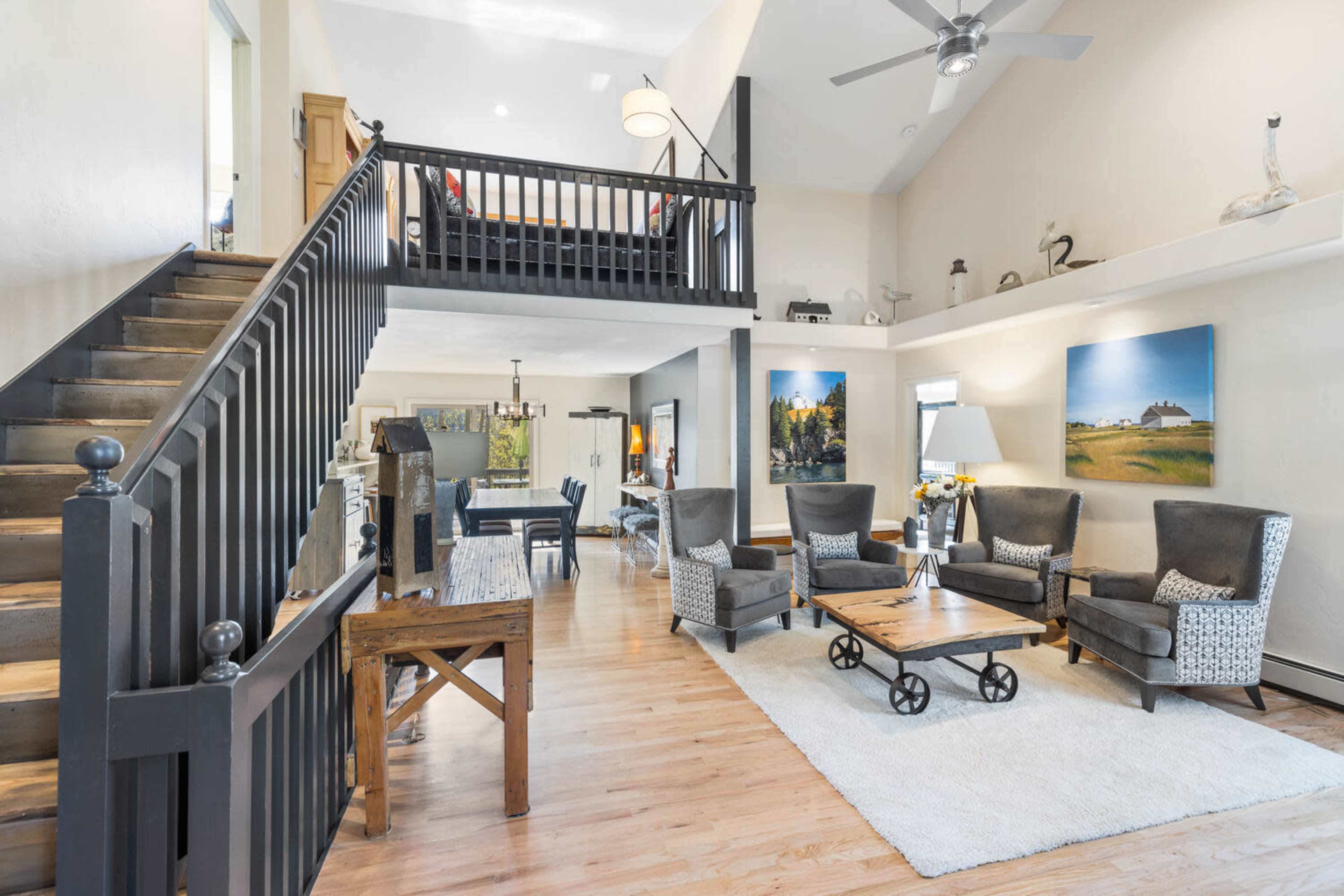 The image shows a spacious living room with a staircase leading to an upper loft area, featuring modern furniture arranged around a central coffee table on a rug.