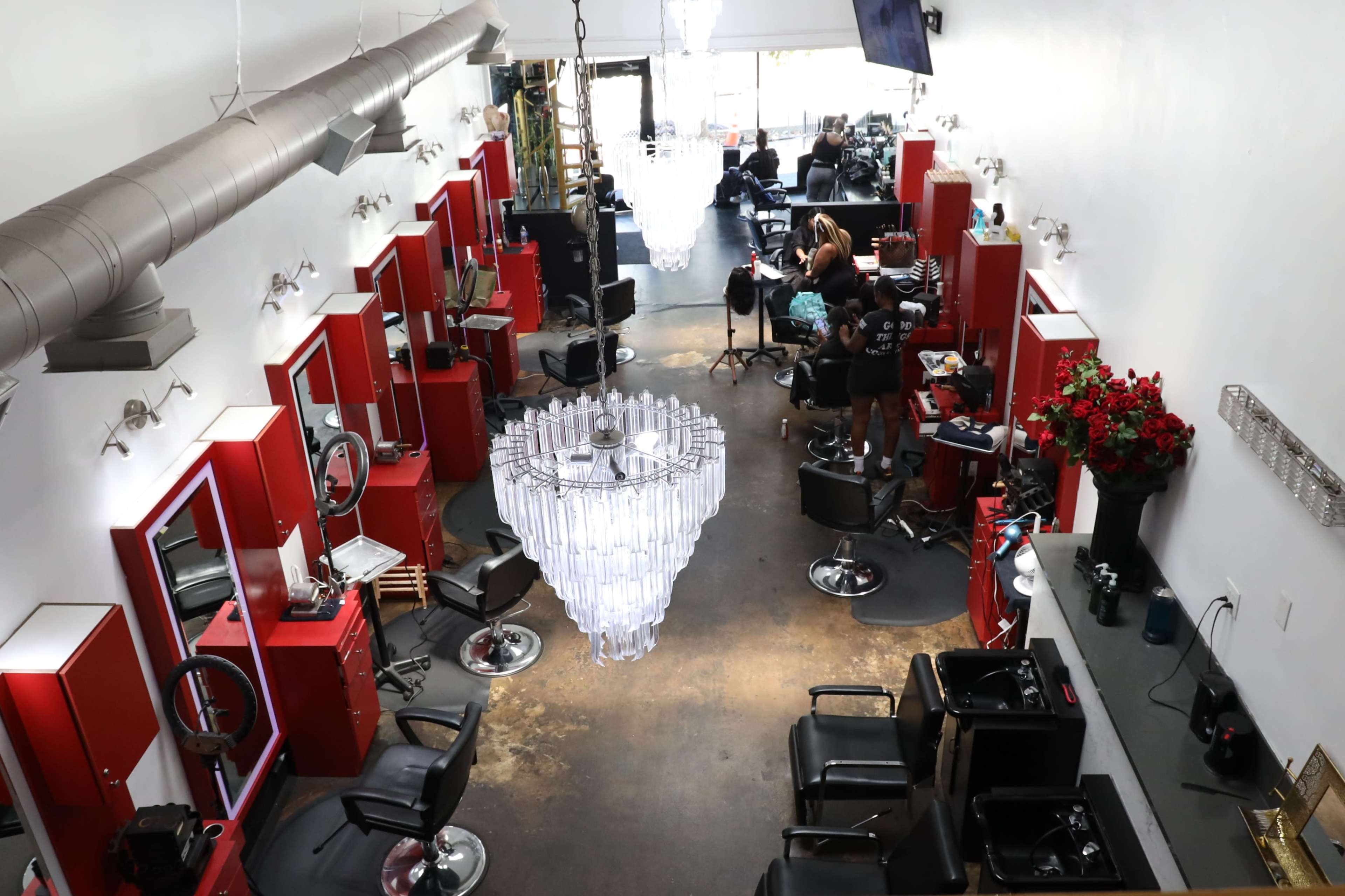 A hair salon with red styling stations, a large chandelier in the center, and stylists working on clients.