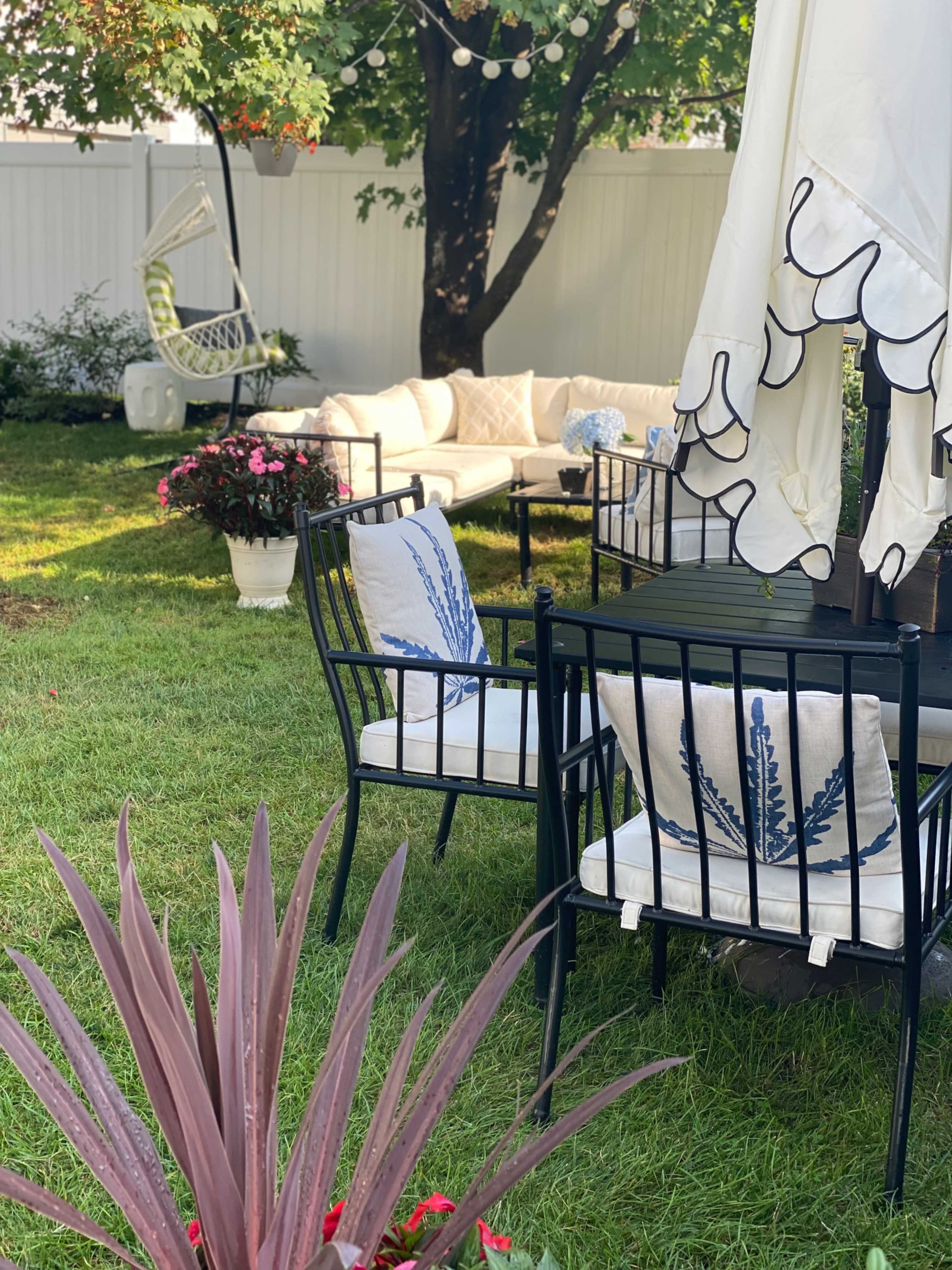 A landscaped backyard featuring a seating area with black metal chairs and a white patio umbrella, surrounded by decorative plants and a white couch.