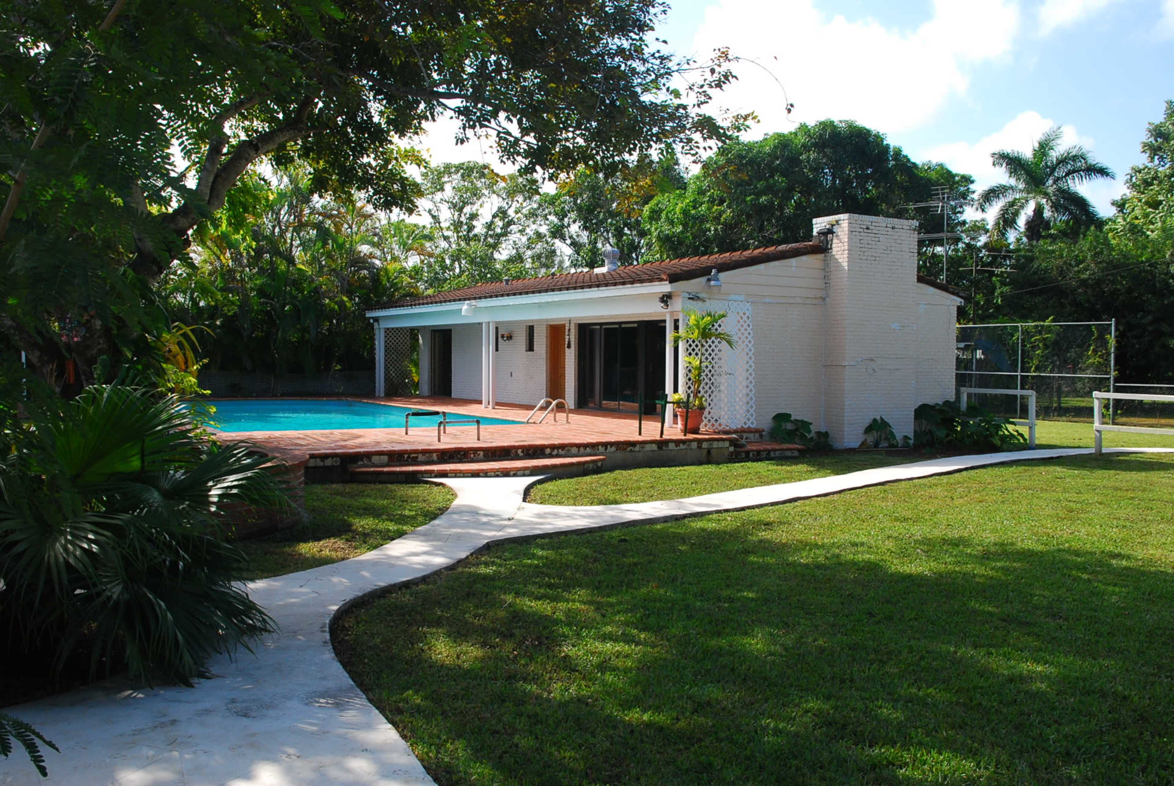A single-story house with a pool and a paved walkway surrounded by grass and trees.