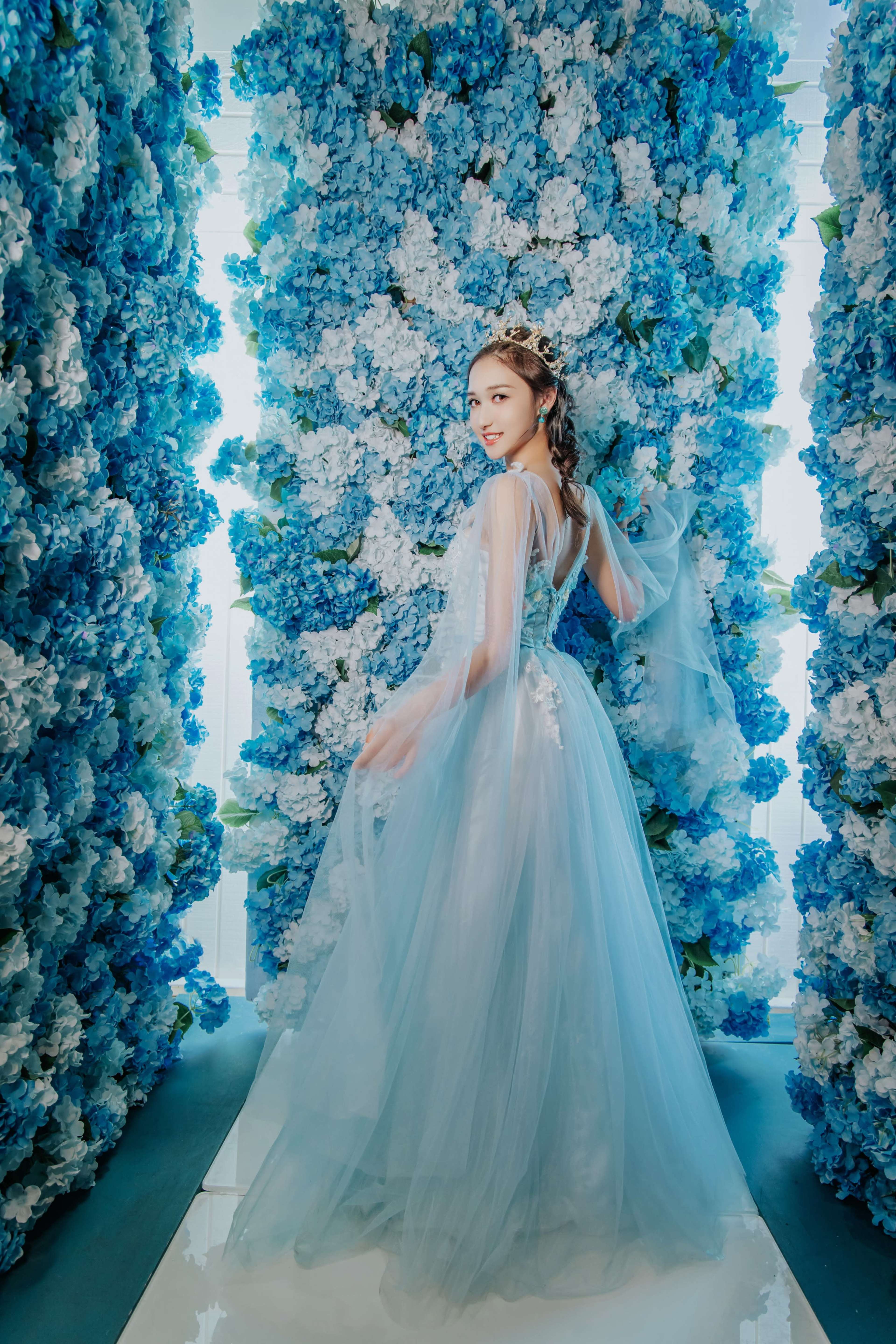 A young woman in a flowing gown poses in a corridor surrounded by vibrant blue and white floral walls.