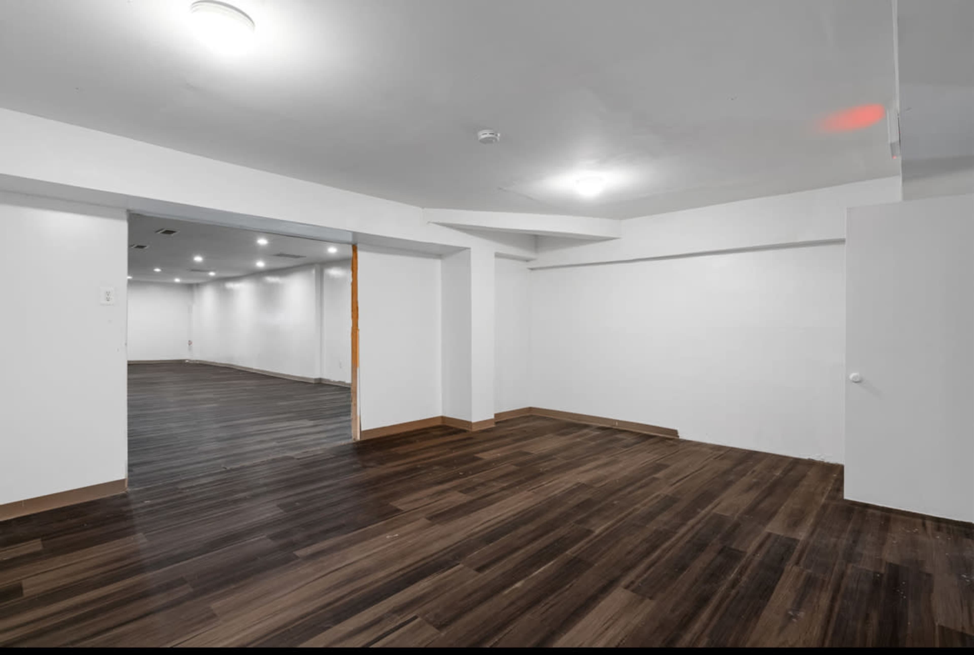 The image shows an empty, well-lit room with dark wooden flooring and white walls, featuring two entrances and a corner area.