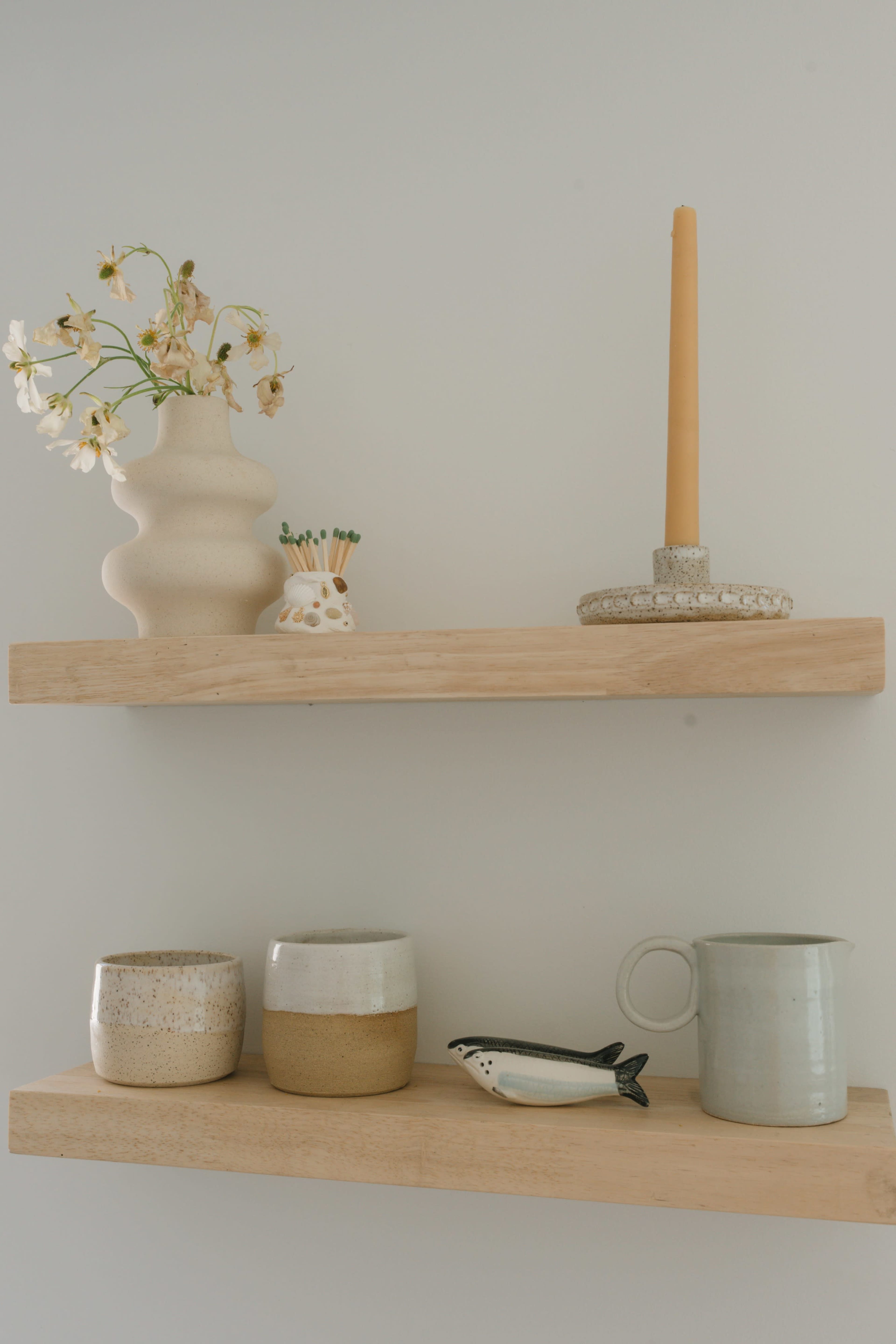The image shows a wooden shelf displaying a decorative vase with flowers, a candle holder with a lit candle, and several ceramic containers along with a small fish figurine and a pitcher.