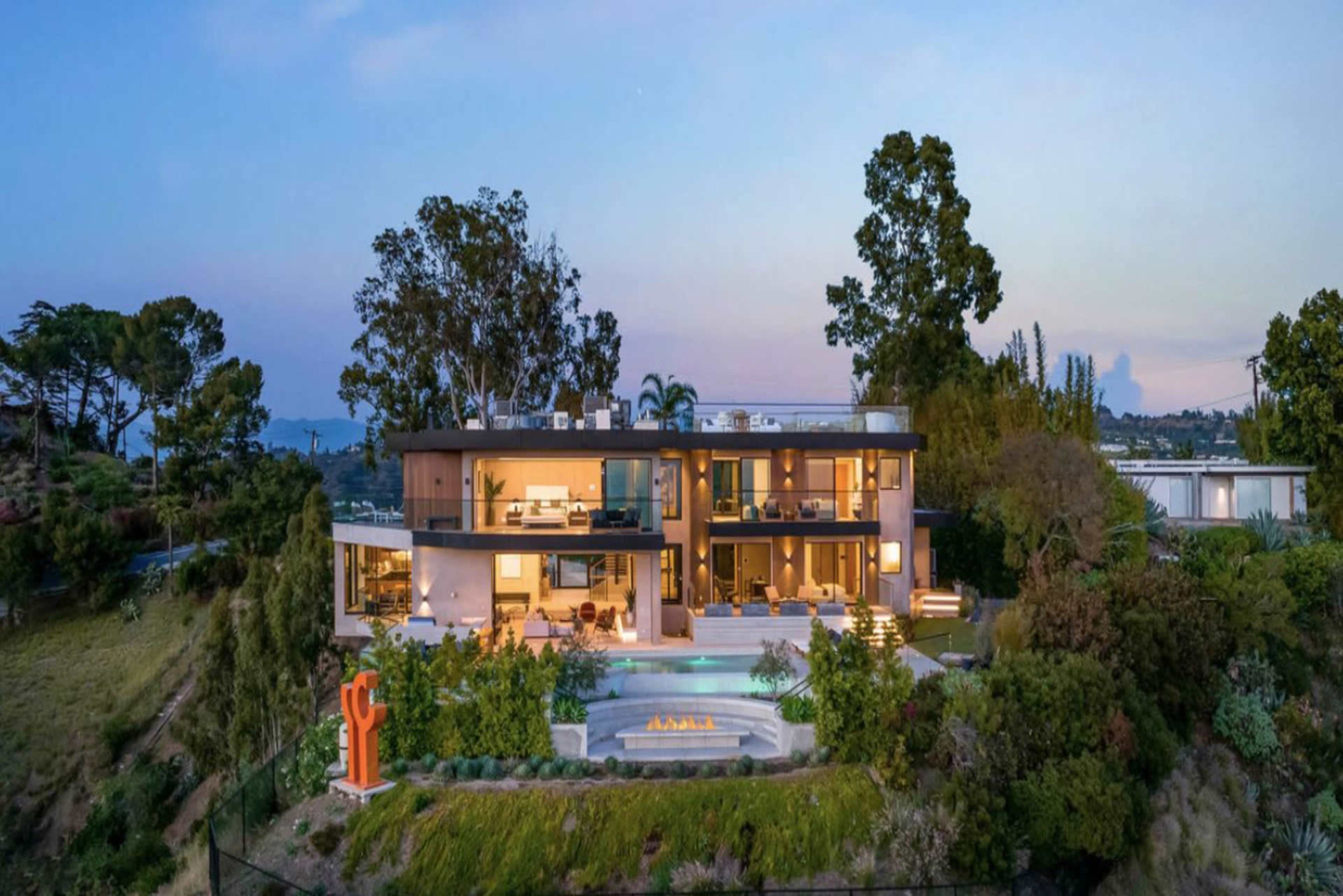 A modern two-story house, featuring large windows and a pool, is set on a hillside surrounded by trees and landscaping.