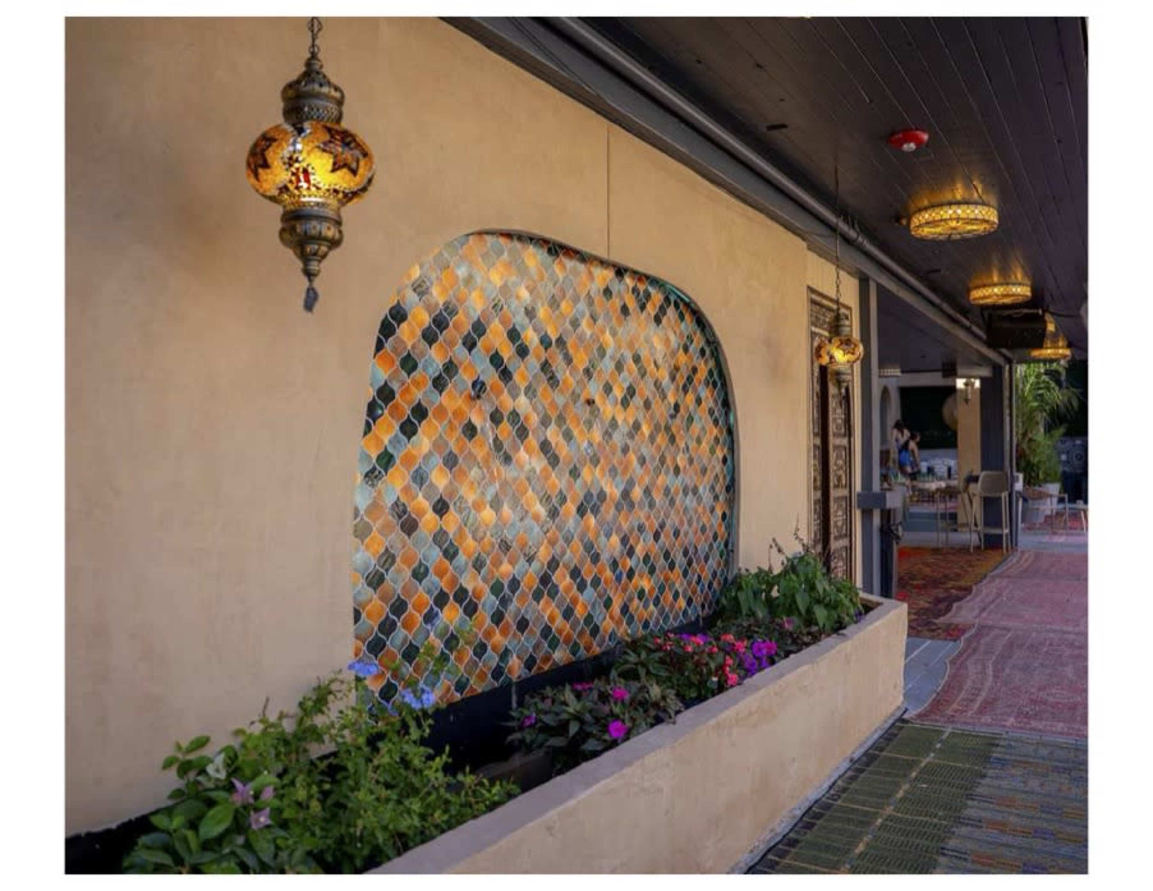 The scene features a textured wall adorned with colorful tiles and overhead lanterns, accompanied by a planter filled with flowers.