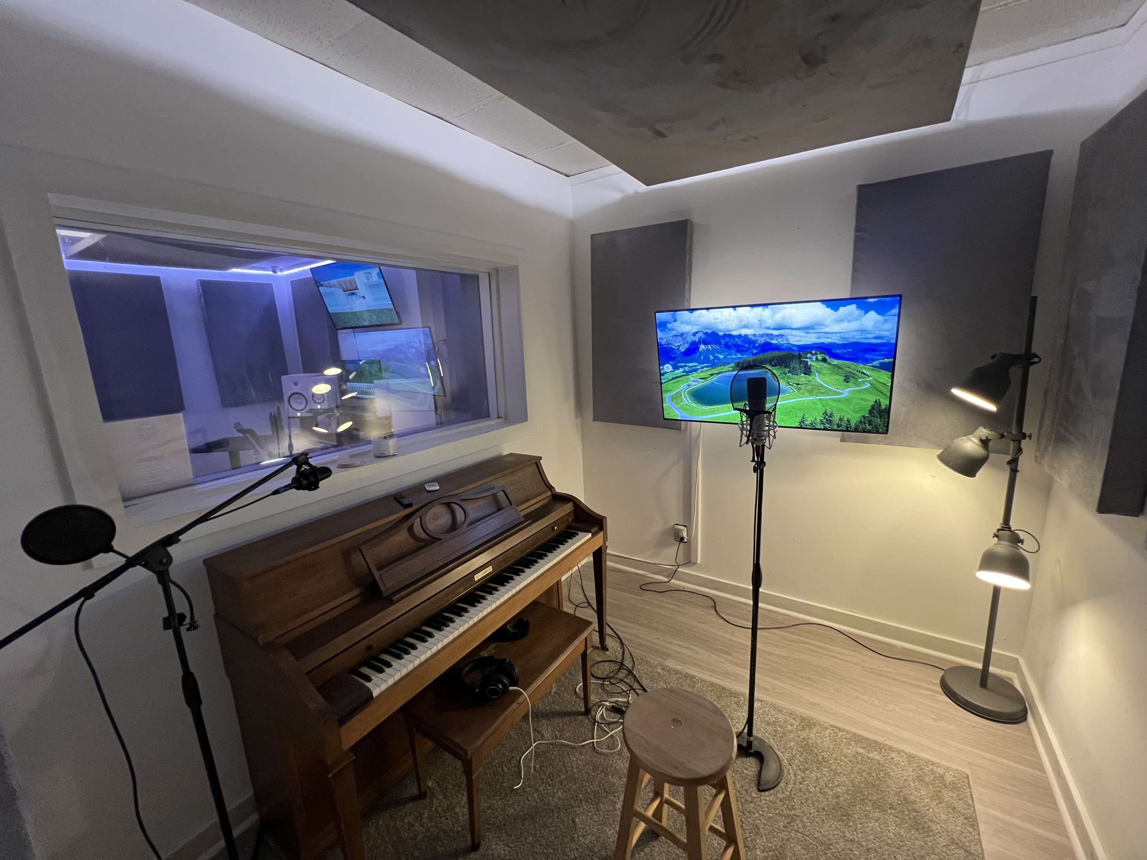 A small recording studio with a piano, microphones, a stool, and a television displaying a landscape scene.