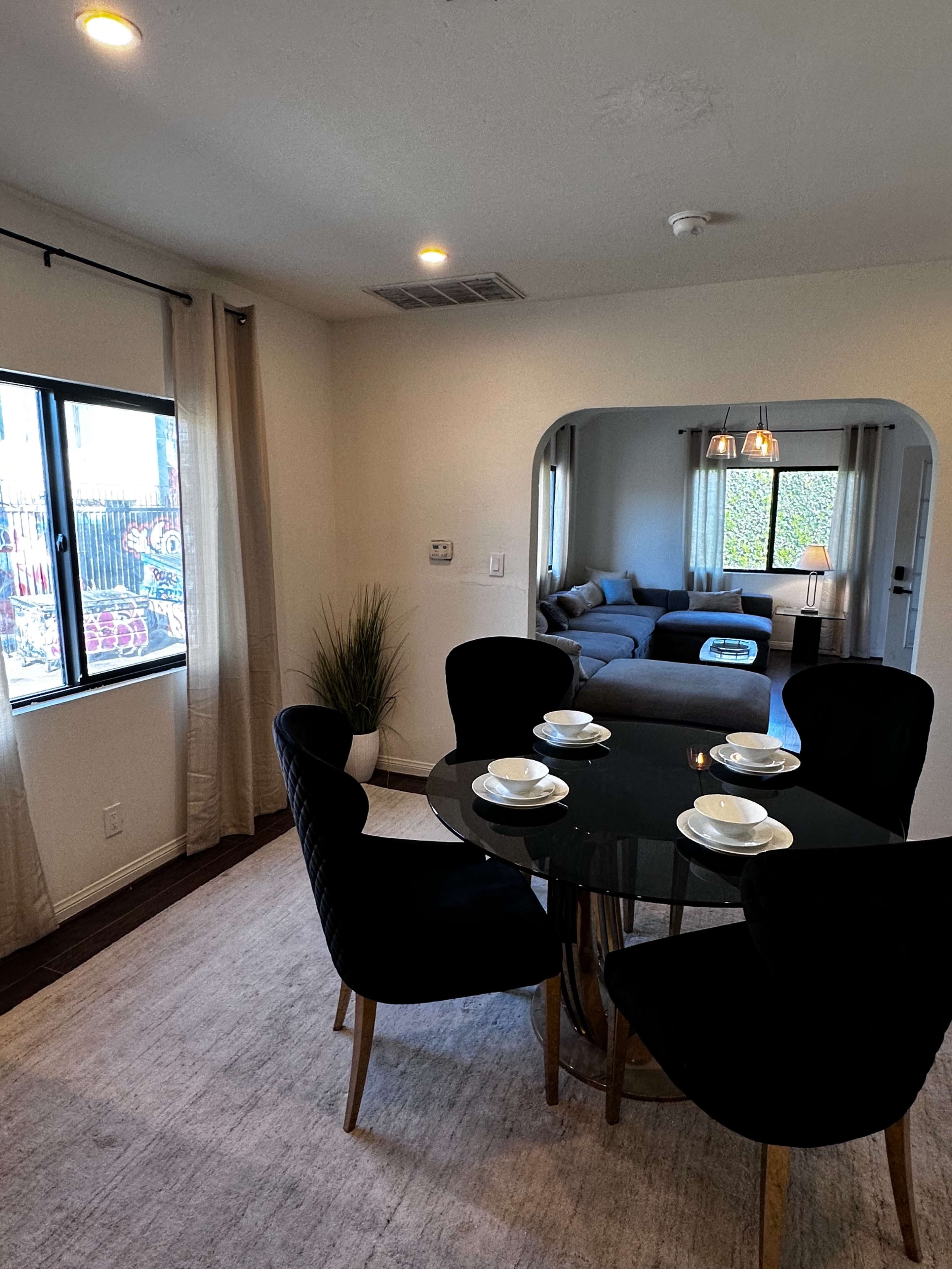 A dining area features a round black table set with white cups and saucers, surrounded by four black chairs, with a view of a living room in the background.