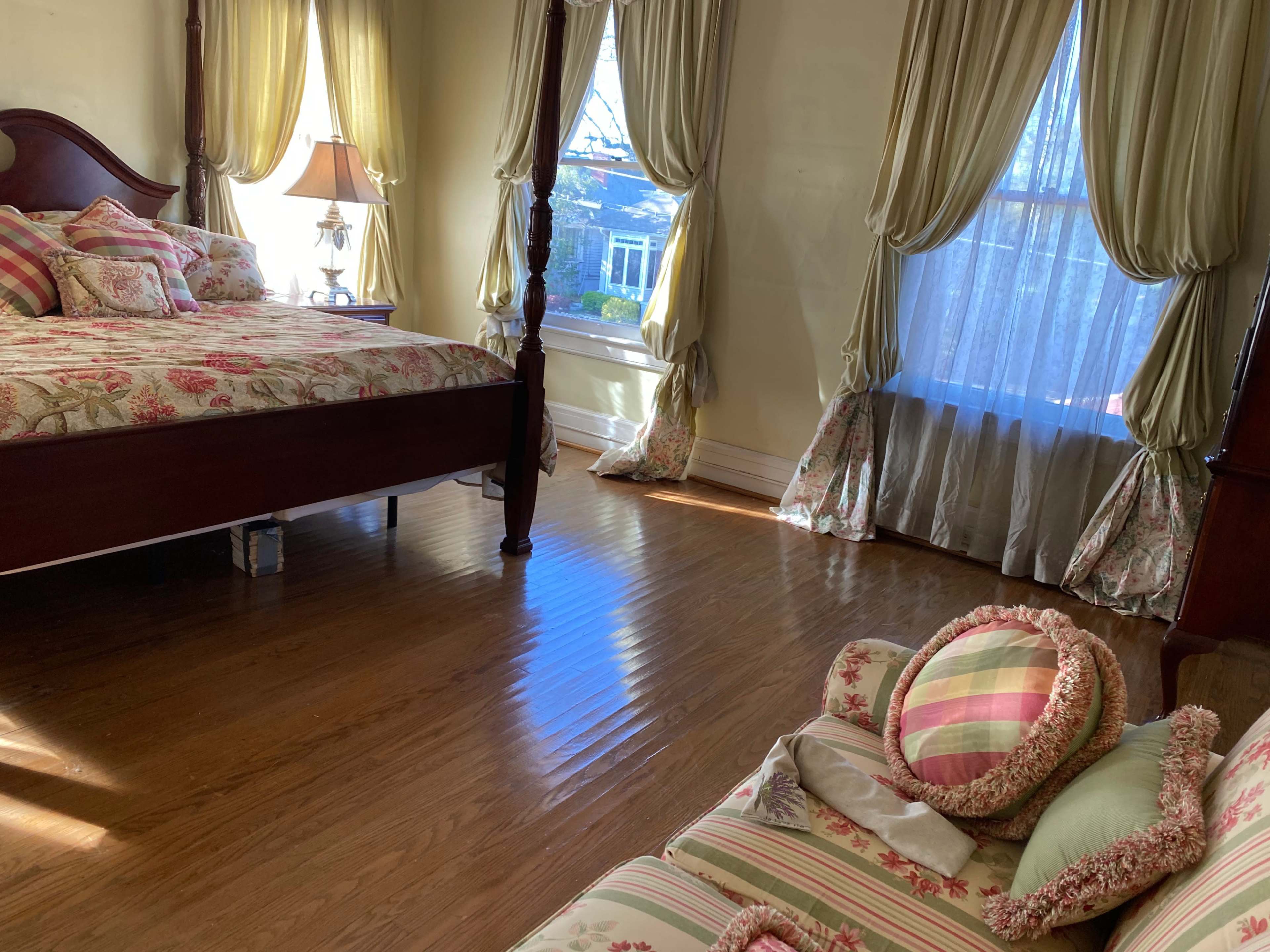 A bedroom features a large bed with floral bedding, a lamp on a nightstand, and light streaming through sheer curtains.