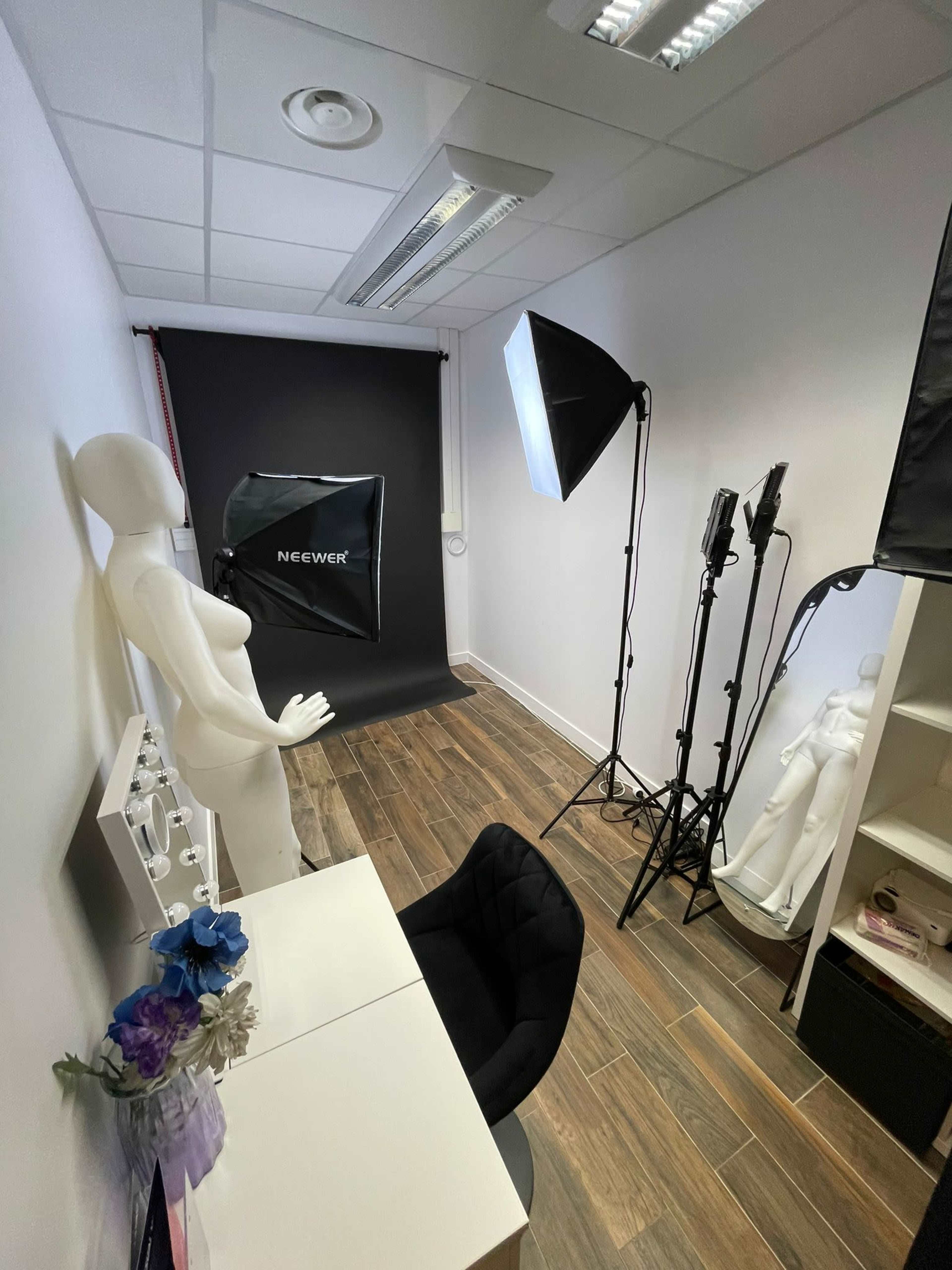 A small studio space featuring a white mannequin, a black backdrop, photography lighting equipment, and a desk with a chair.