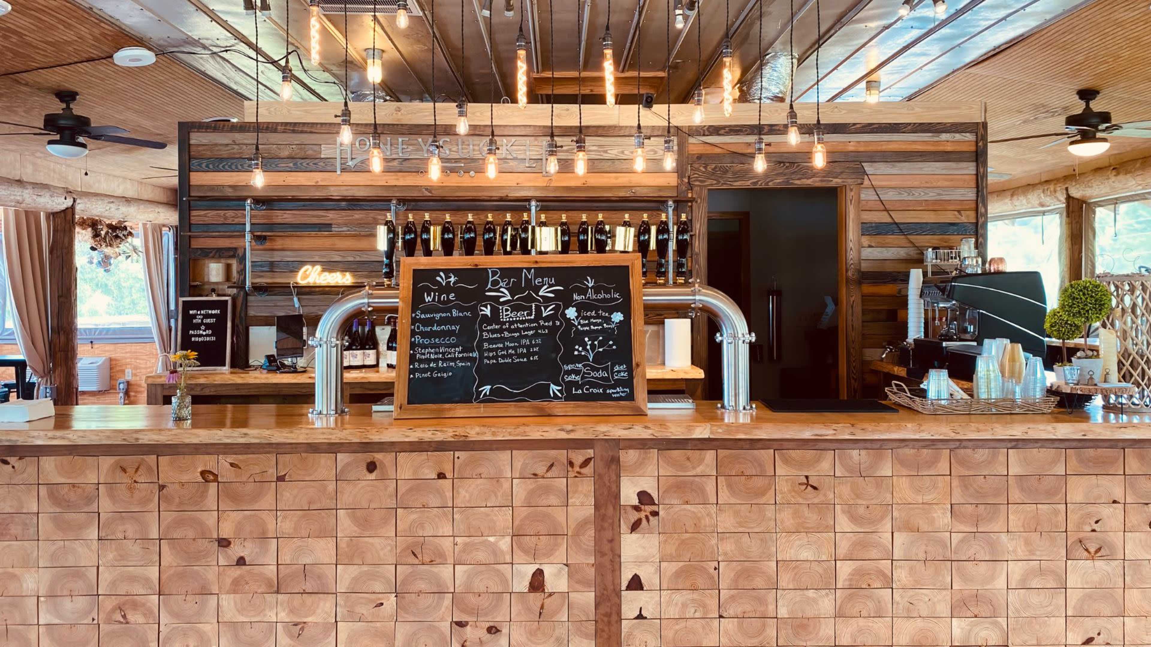A bar with a wooden counter displays a chalkboard menu surrounded by wine bottles and pendant lights hanging from the ceiling.