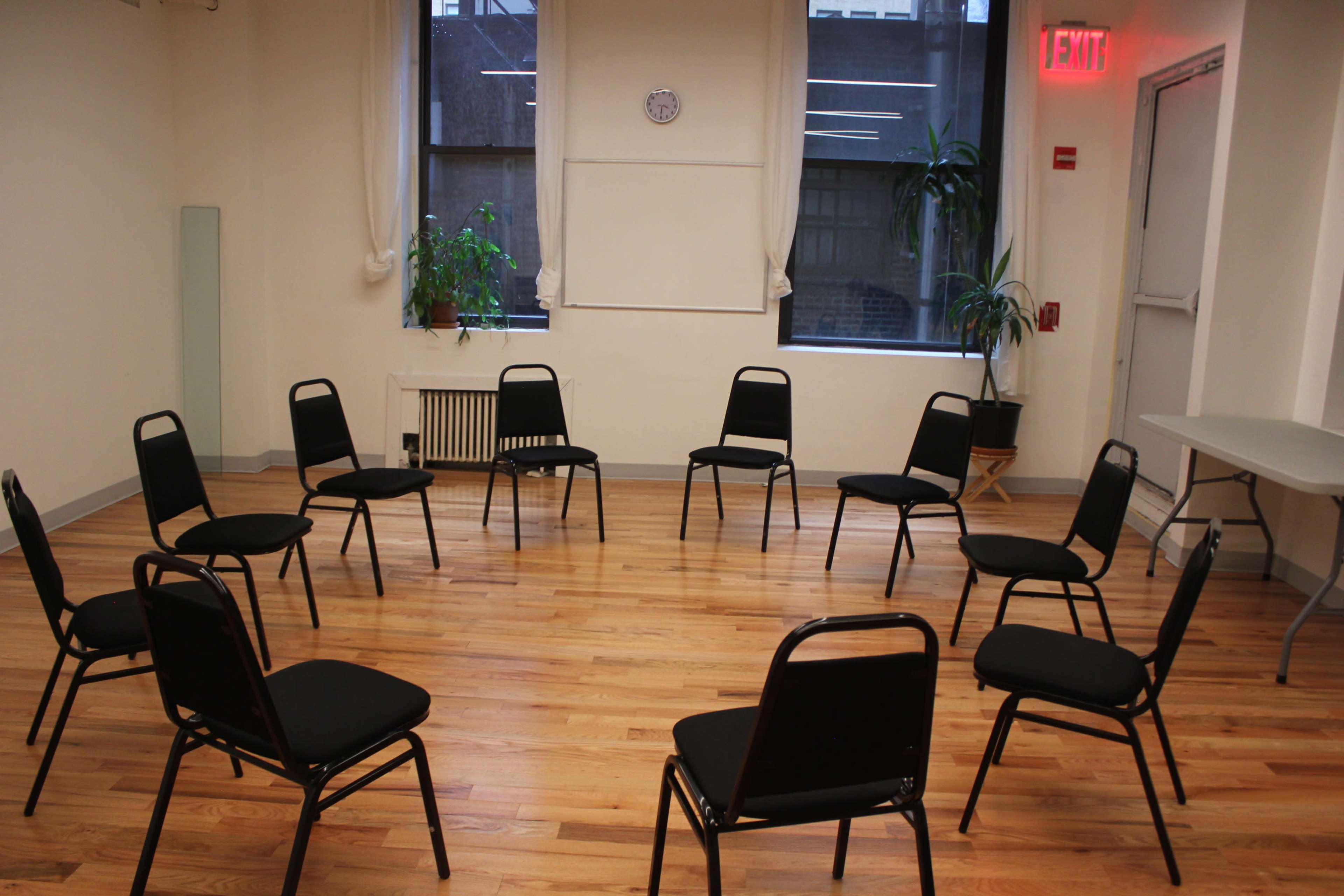 A room with hardwood floors arranged with twelve black chairs in a circle, two windows allowing natural light, and a small table against one wall.