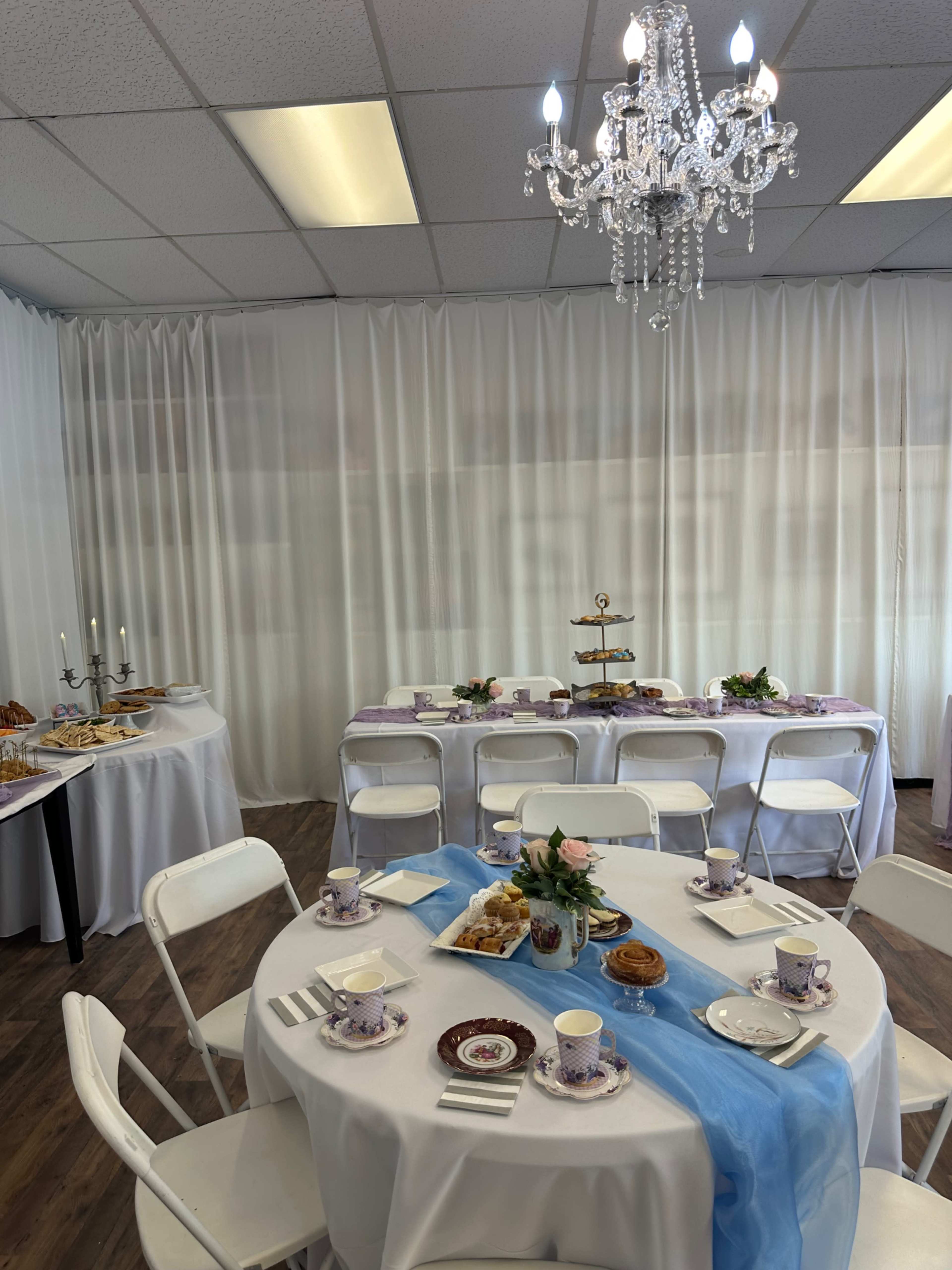 A decorated room features a round table set with tea cups and pastries, surrounded by white chairs, with a buffet table and a chandelier overhead.