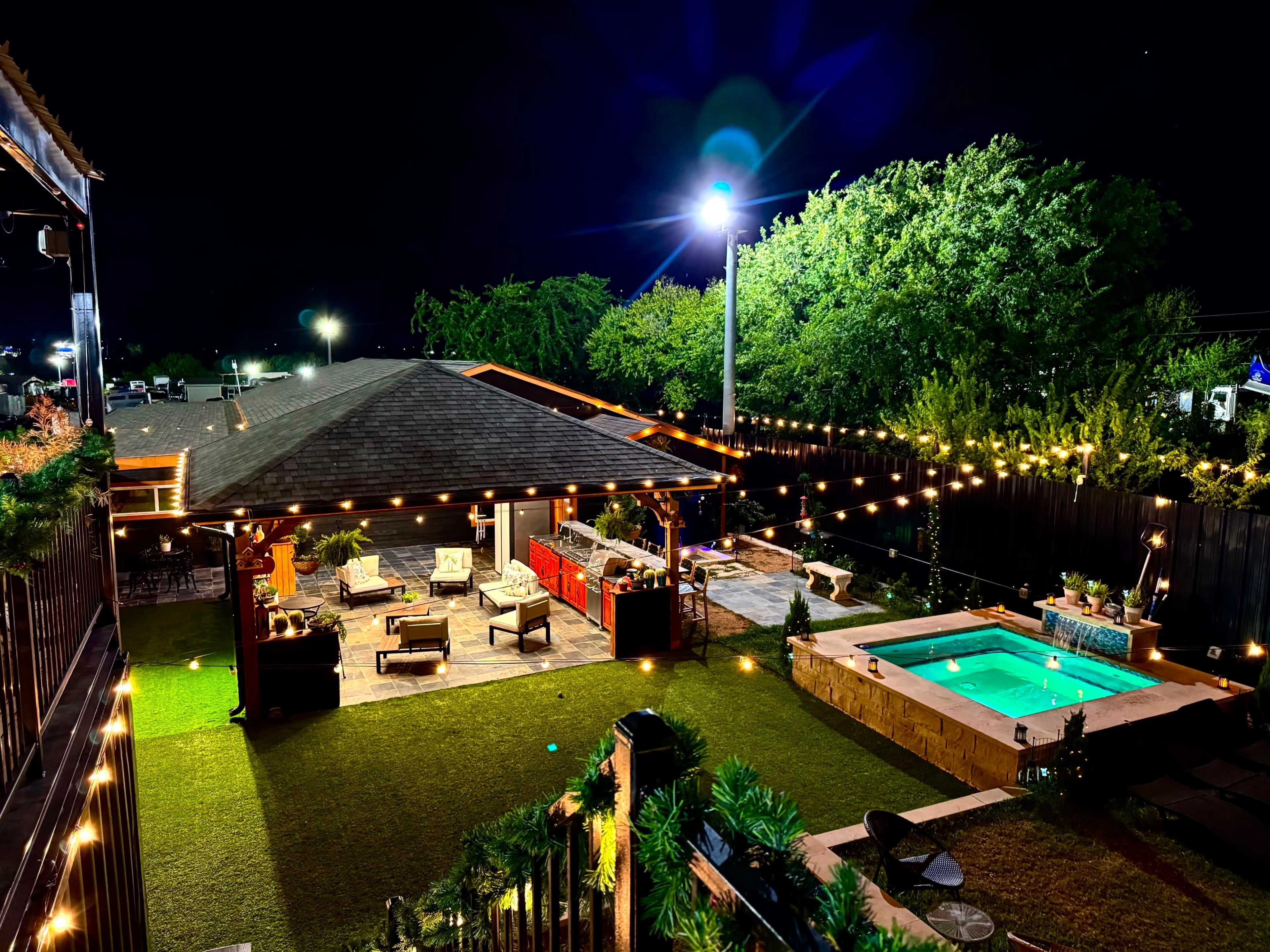 The image shows a well-lit backyard at night featuring a patio with seating, an outdoor kitchen, and a small swimming pool surrounded by greenery and string lights.