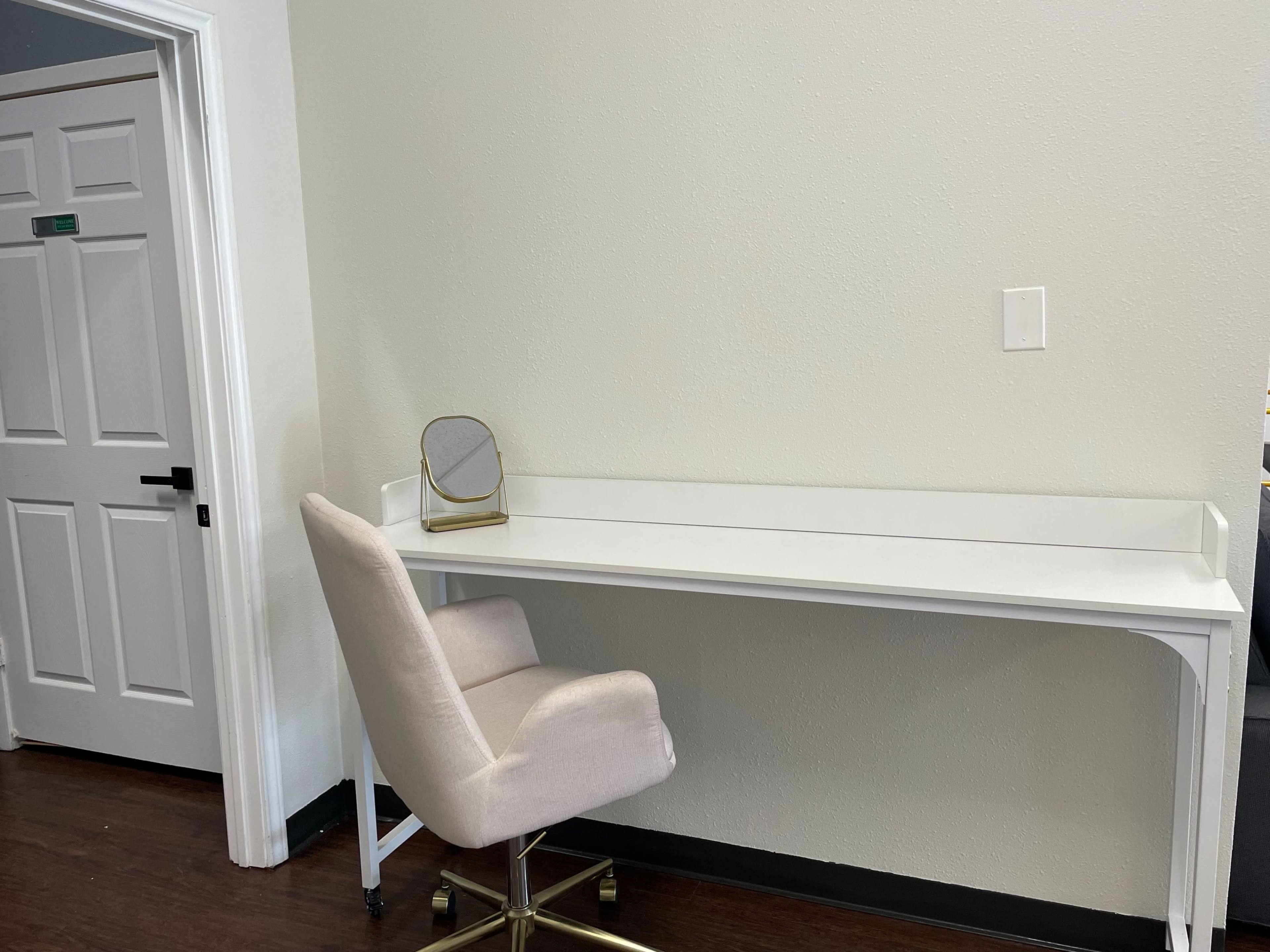 A minimalistic white desk with a mirror sits against a light-colored wall, accompanied by a light-colored chair on wheels.
