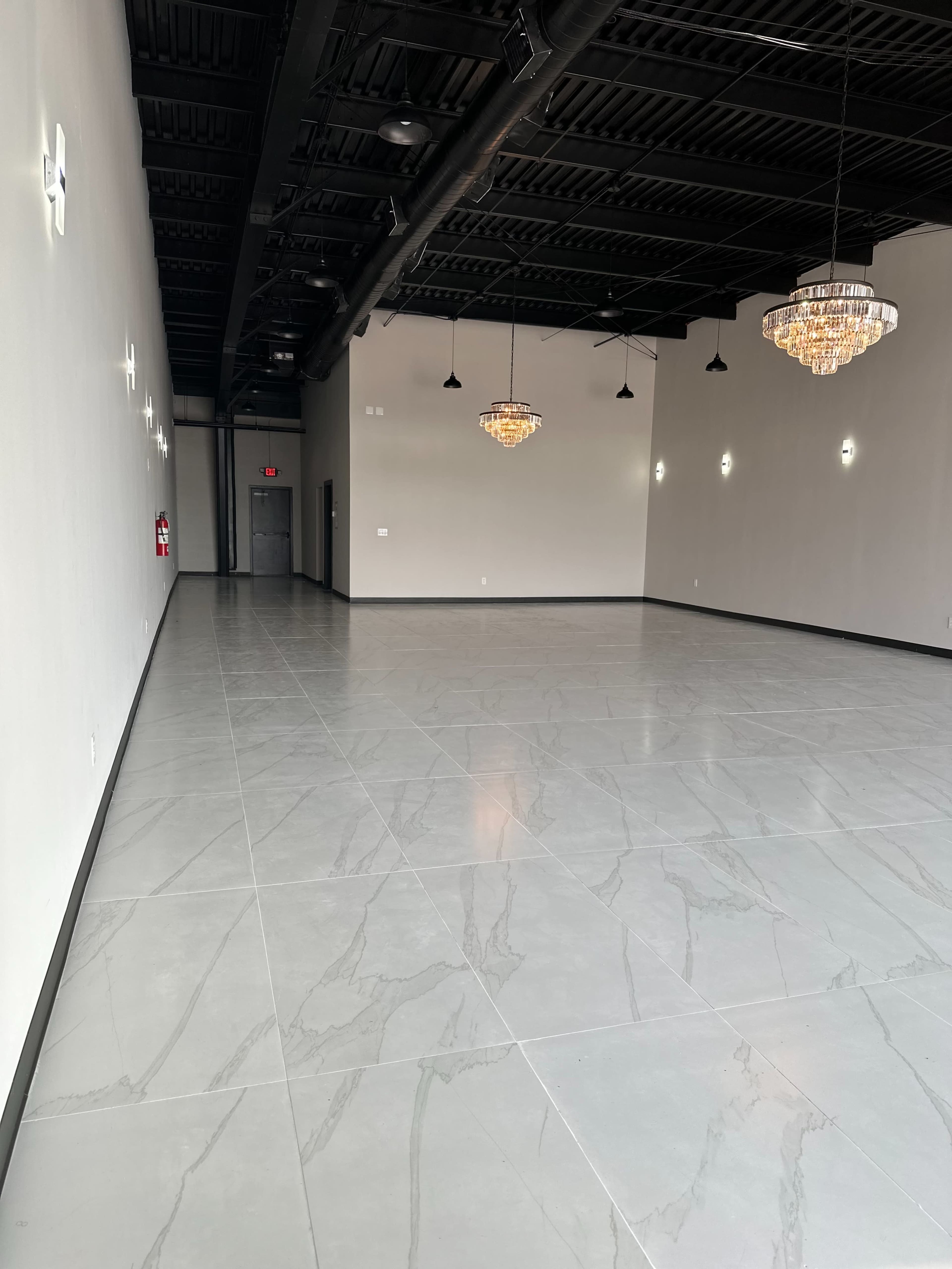 A spacious, empty room with a polished gray floor, tall ceilings, and two crystal chandeliers hanging from the ceiling.