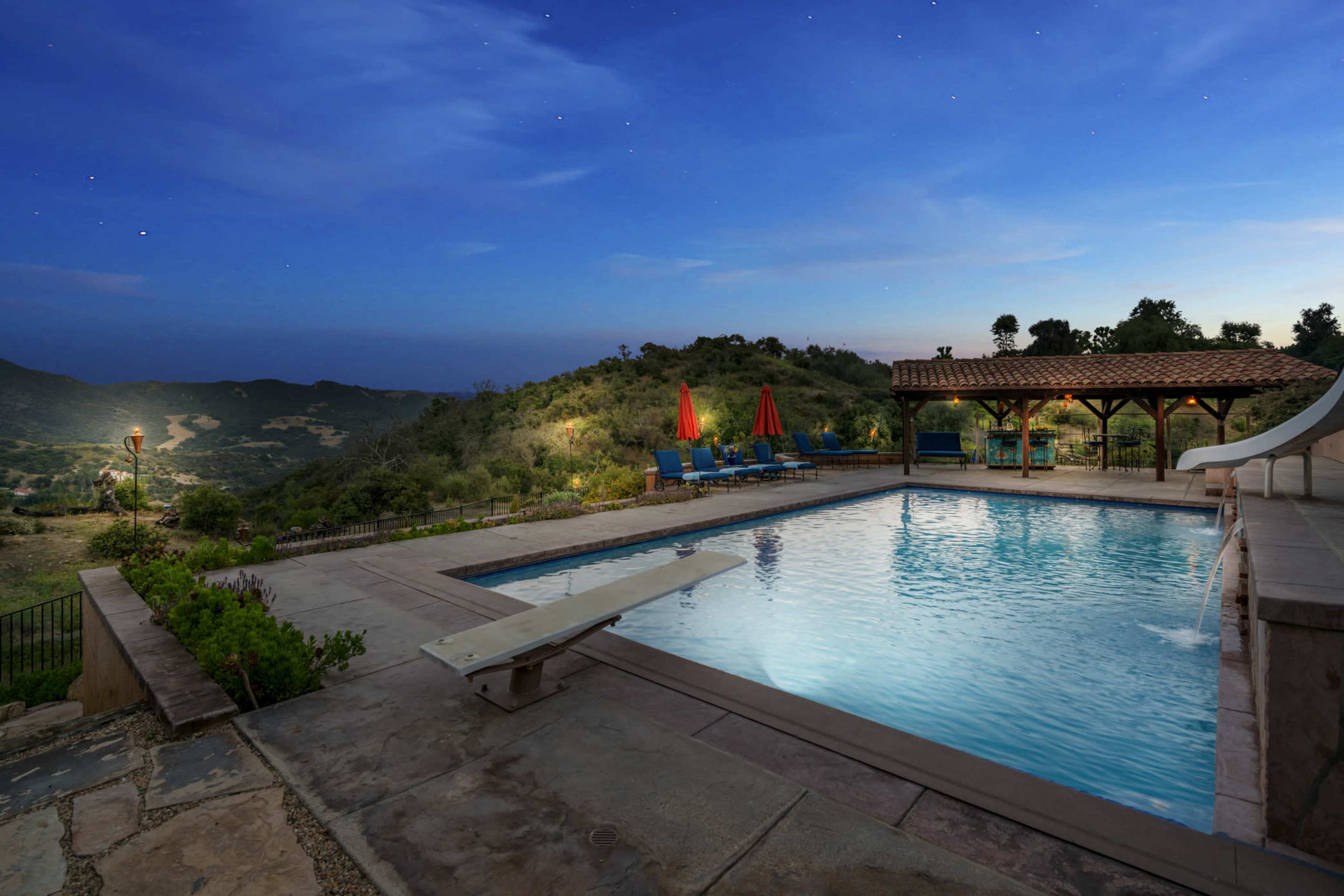 The image shows a pool area surrounded by lush hills, featuring a diving board and shaded seating under a wooden structure with bright red umbrellas.