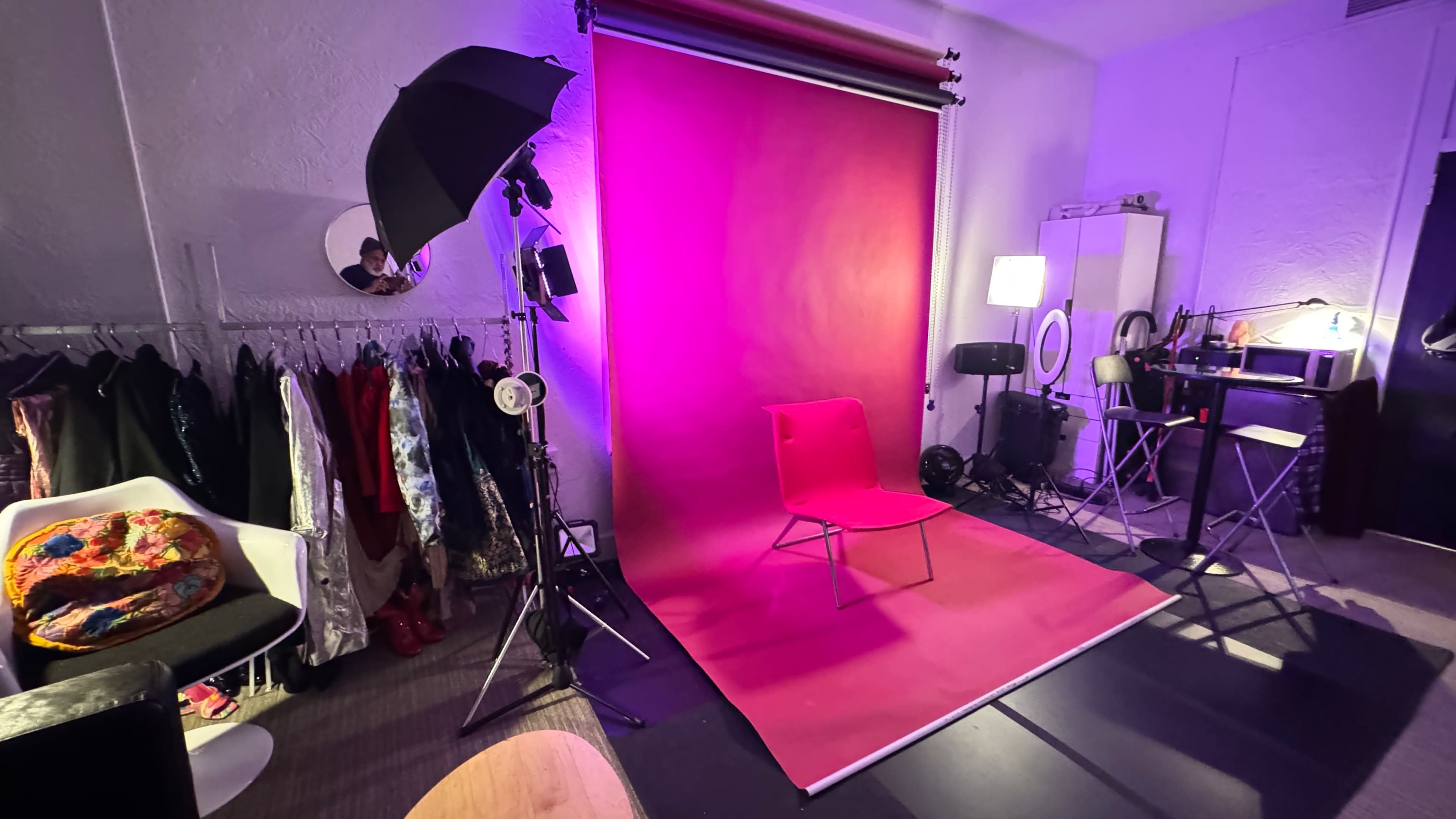 The image shows a studio setup with a bright red backdrop, a chair in the foreground, and various lighting equipment and clothing racks along the sides.