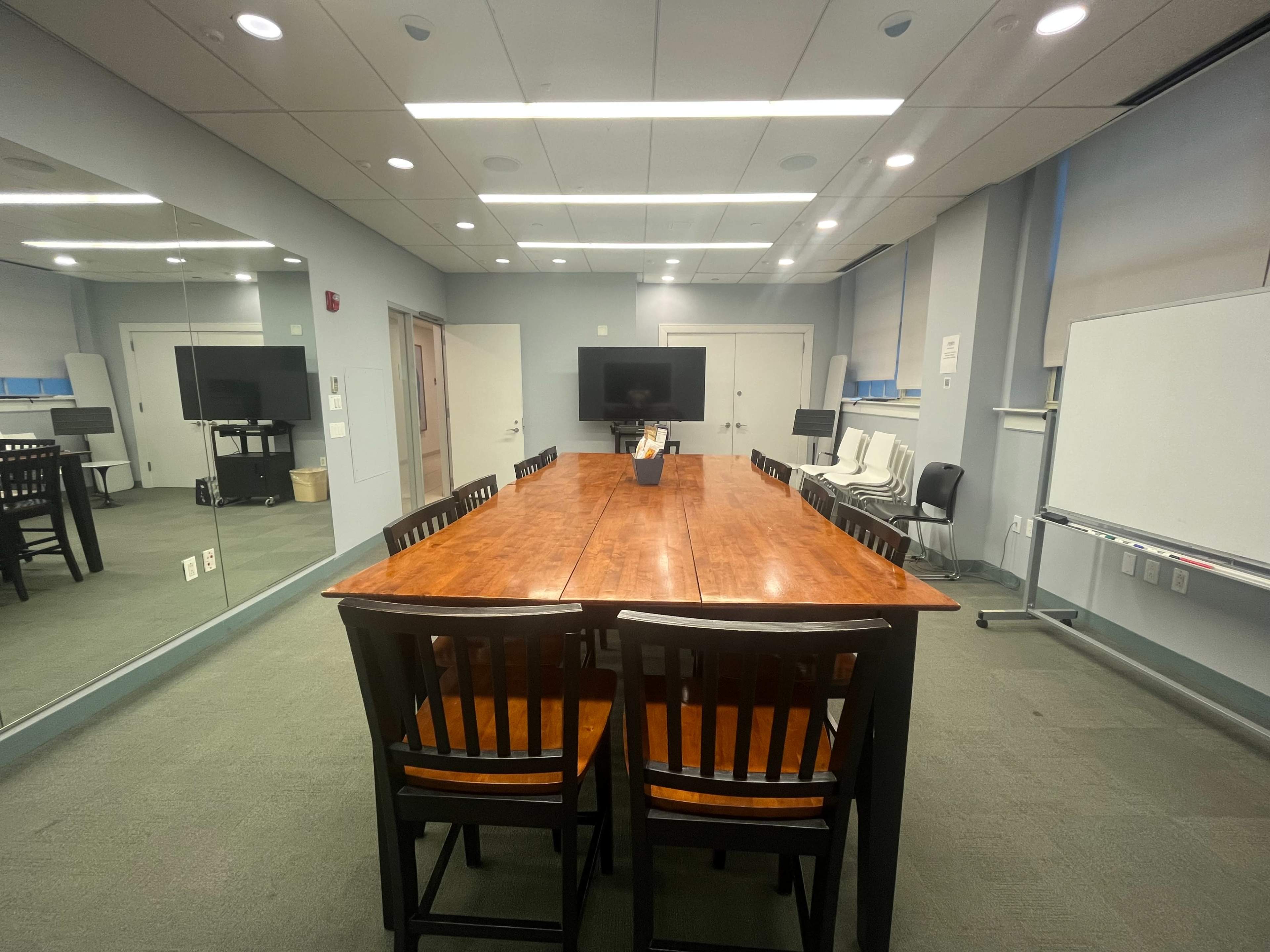 The image shows a spacious conference room with a long wooden table surrounded by chairs, and a large screen at one end.