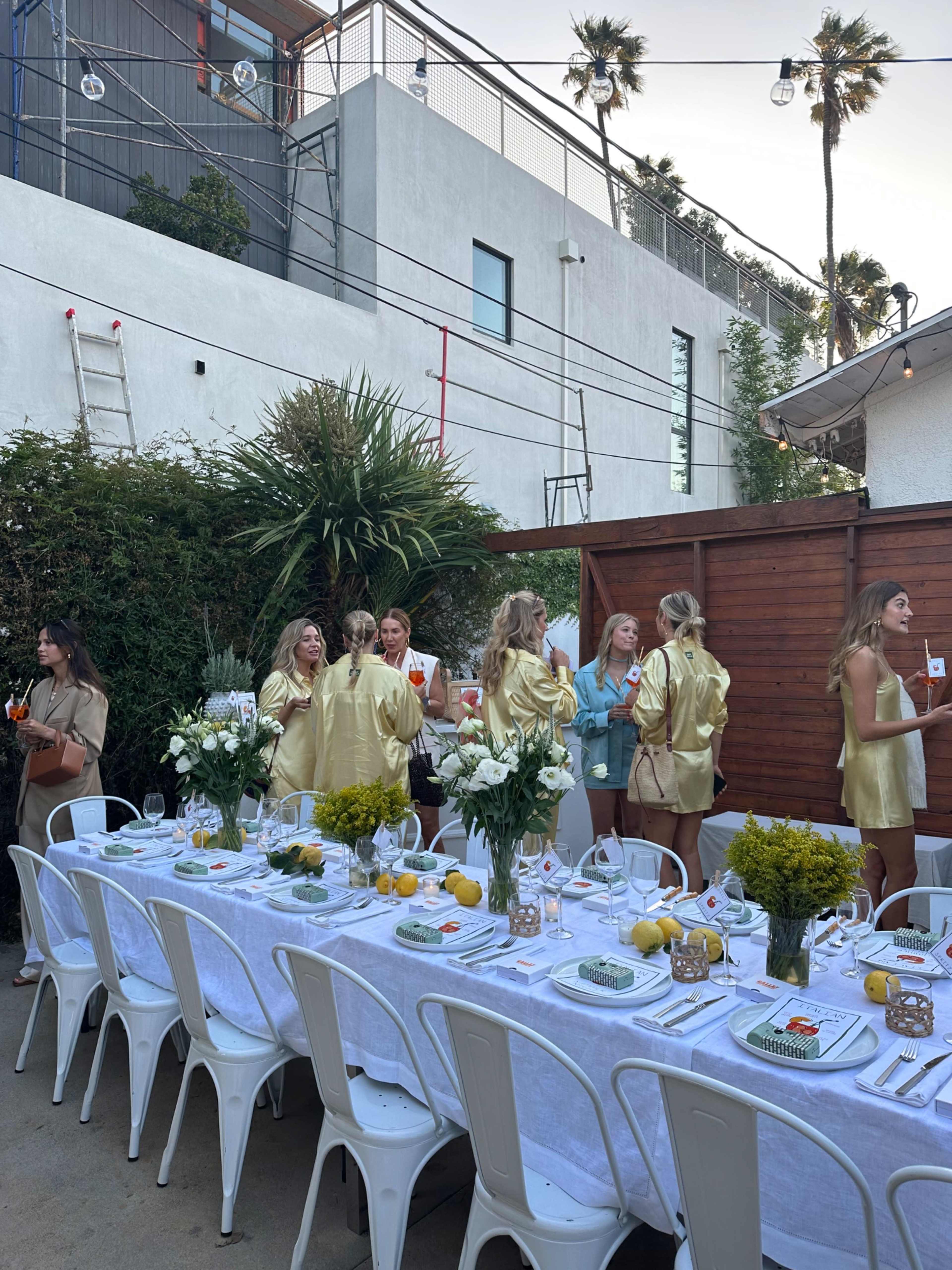 A long dining table is set outdoors with white linens and centerpieces, while a group of women in golden outfits socialize nearby.
