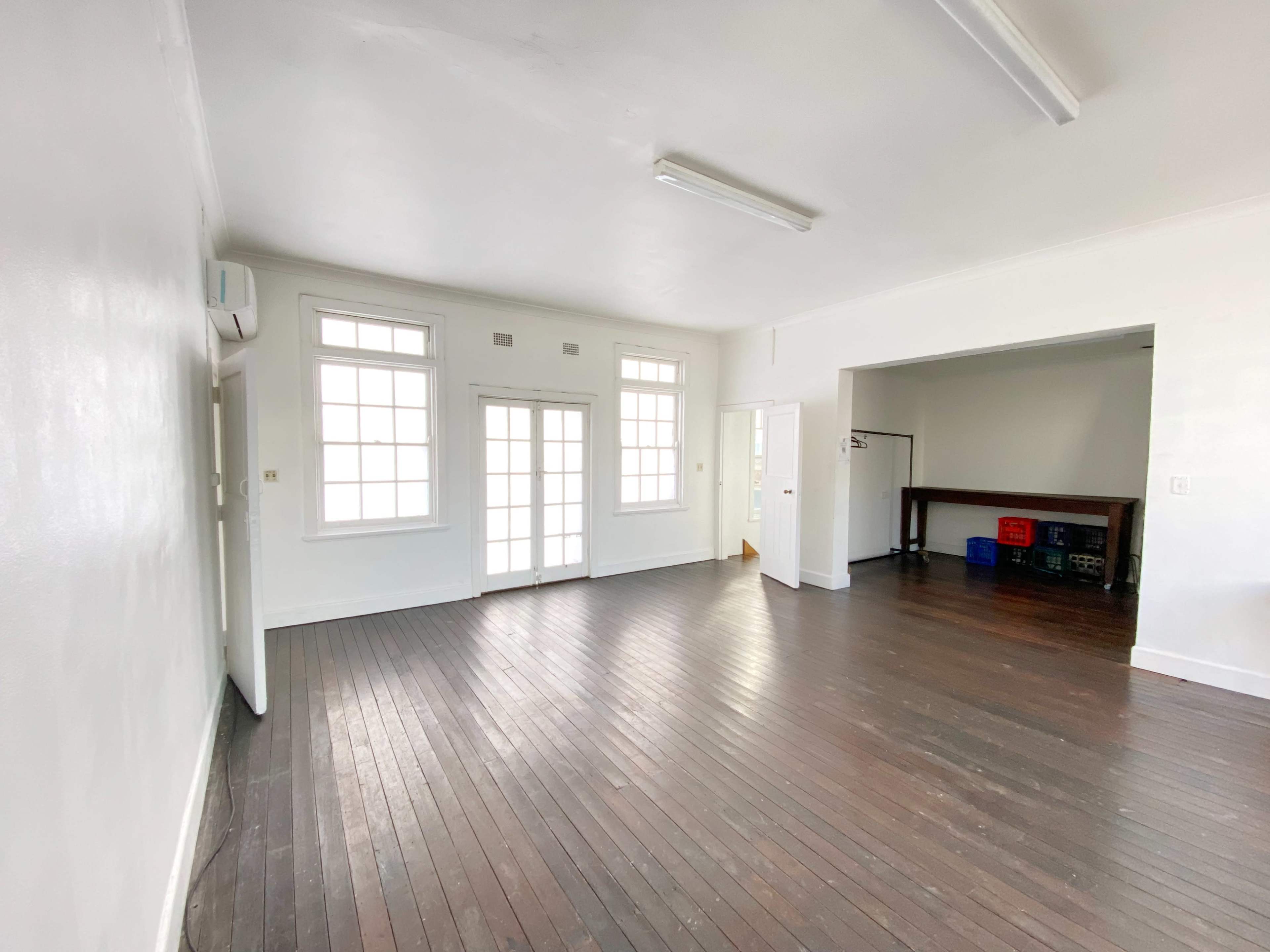 A spacious, empty room with wooden floors and large windows, featuring a table in the adjacent area.