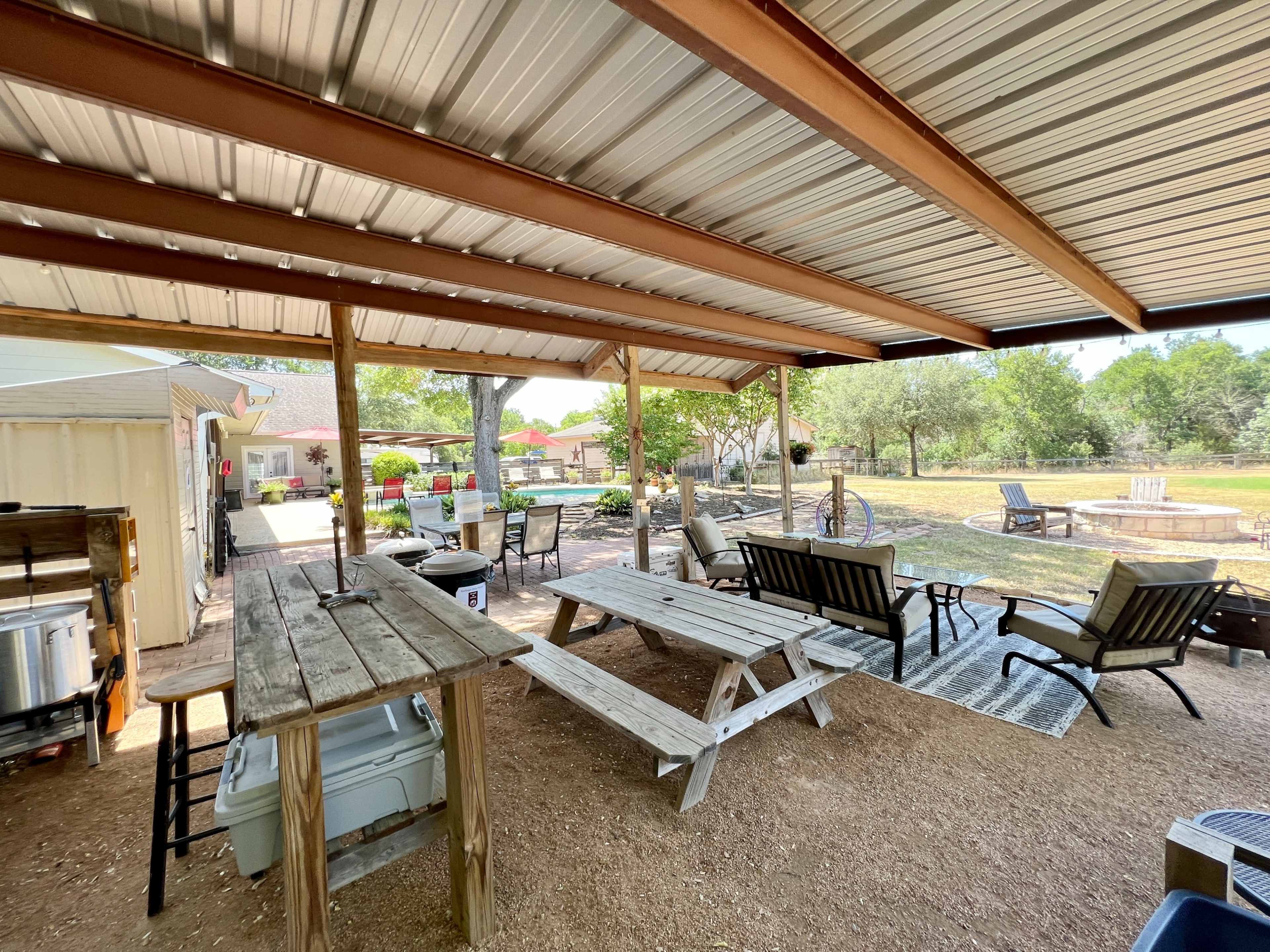 The image shows an outdoor shaded area with wooden picnic tables, lounge chairs, and a view of a landscaped garden.