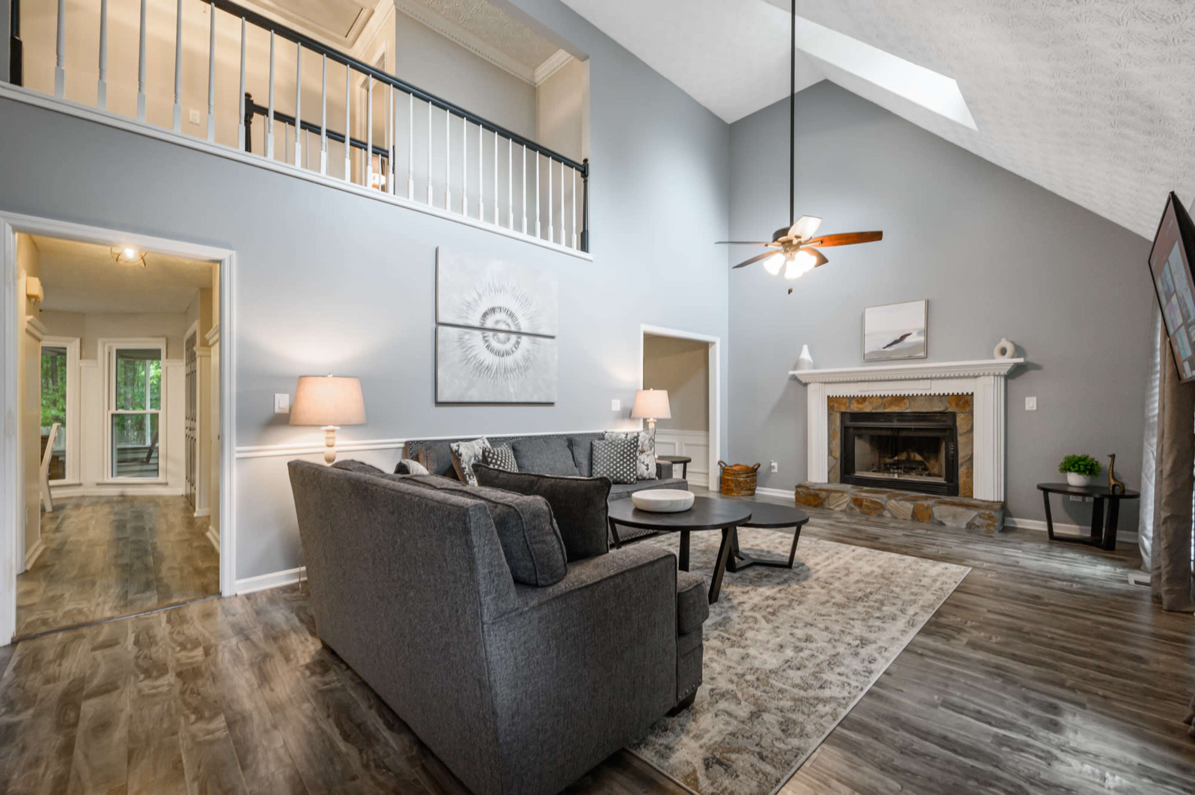 A spacious living room with a high ceiling, a fireplace, and large windows, furnished with a gray sofa and a circular coffee table.