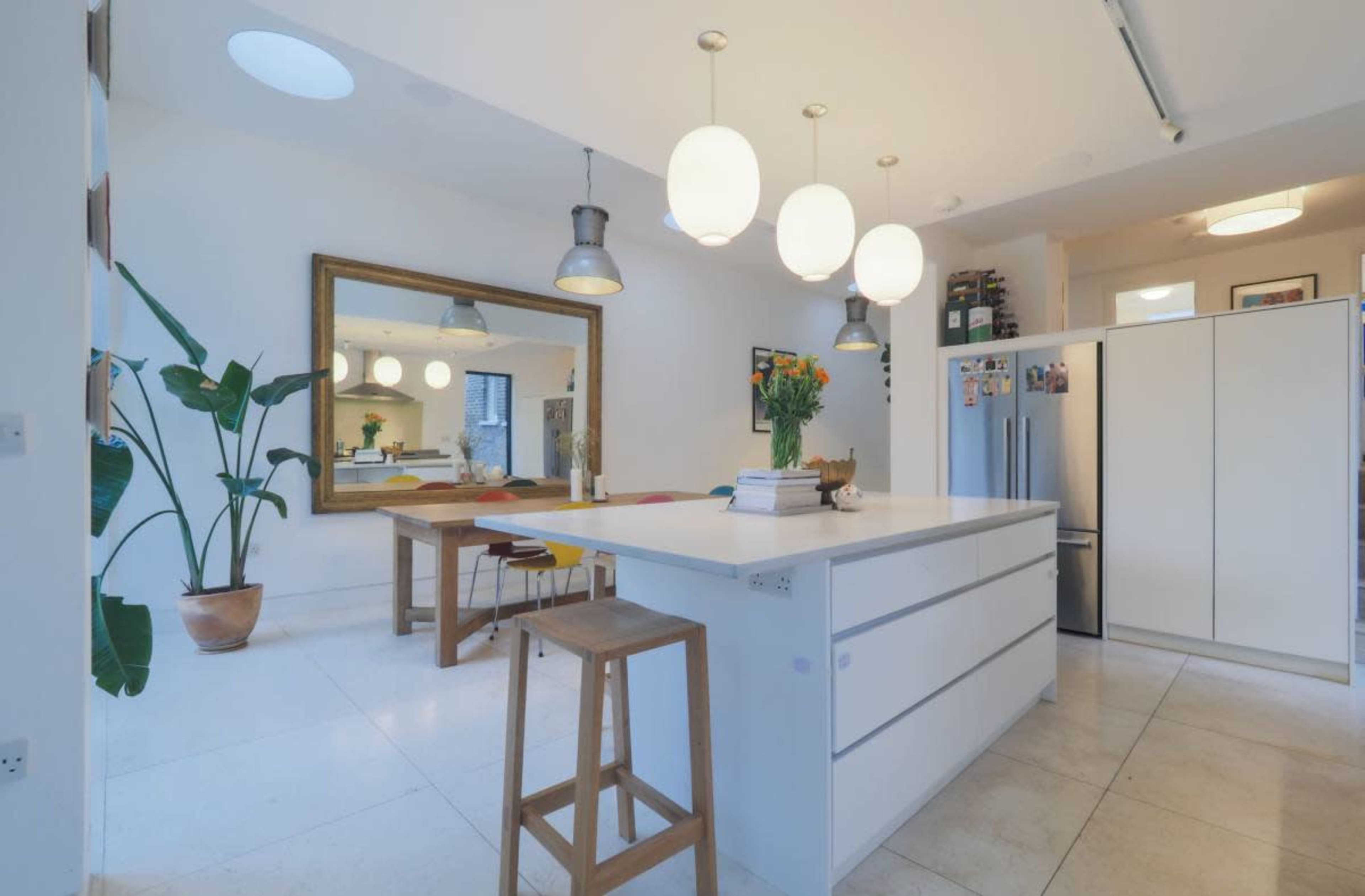 The image shows a modern kitchen with an island, a wooden dining table, and a large mirror reflecting the space, alongside pendant lights and tropical plants.
