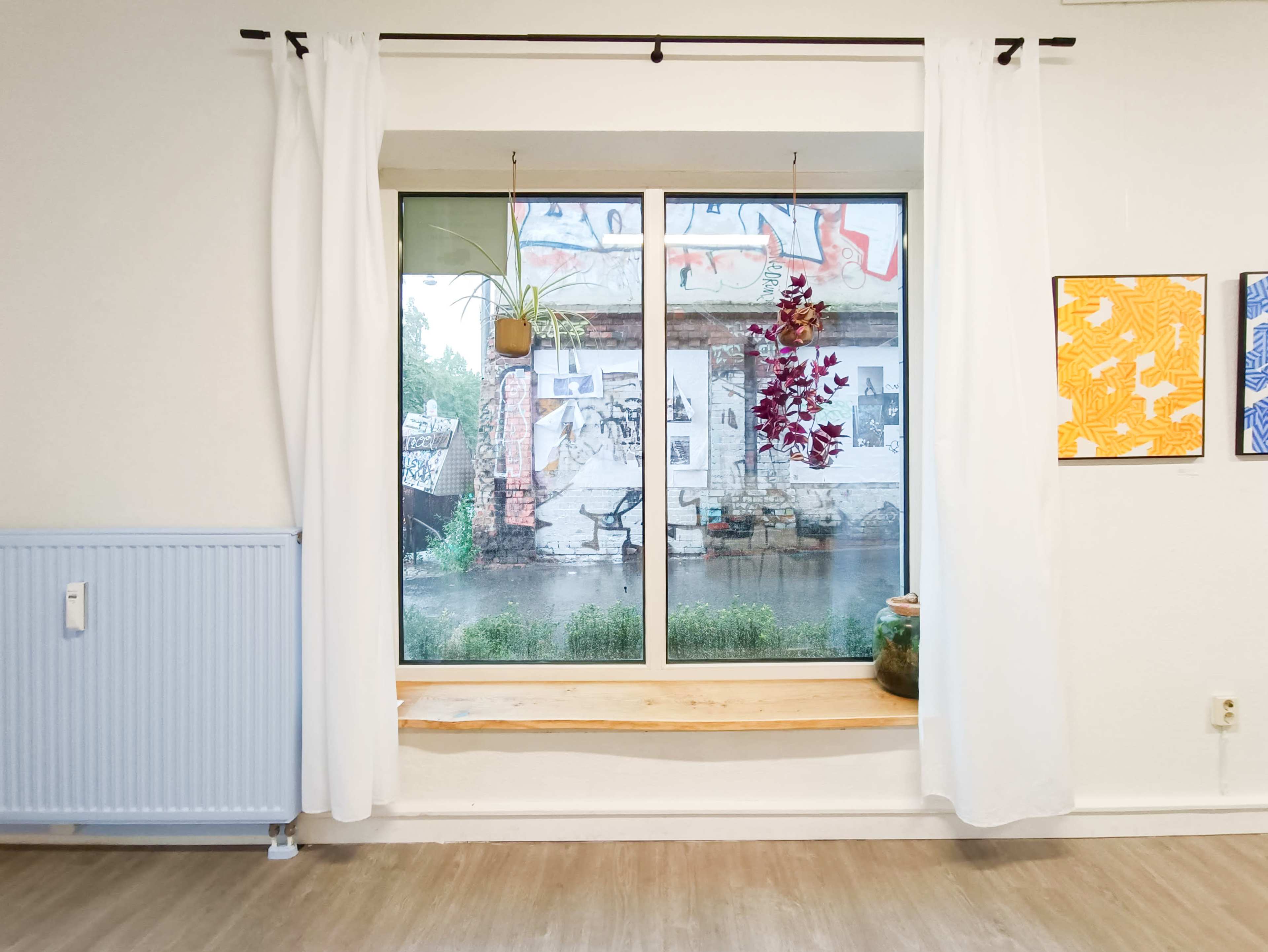 The image shows a large window with white curtains, revealing a rainy outdoor scene with plants and graffiti visible in the background.