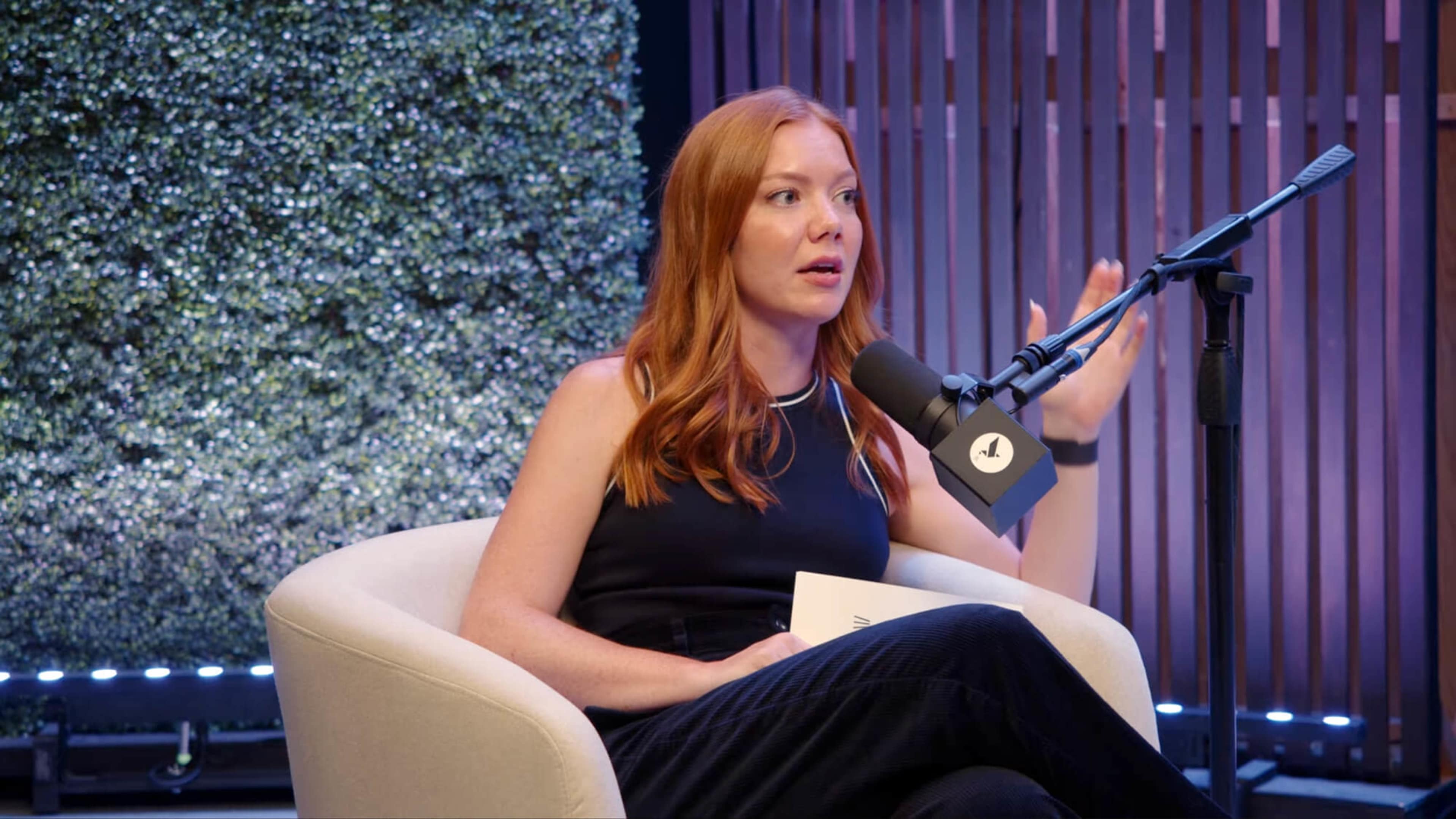 A woman with red hair sits in a cream-colored chair, speaking into a microphone in front of a green backdrop.